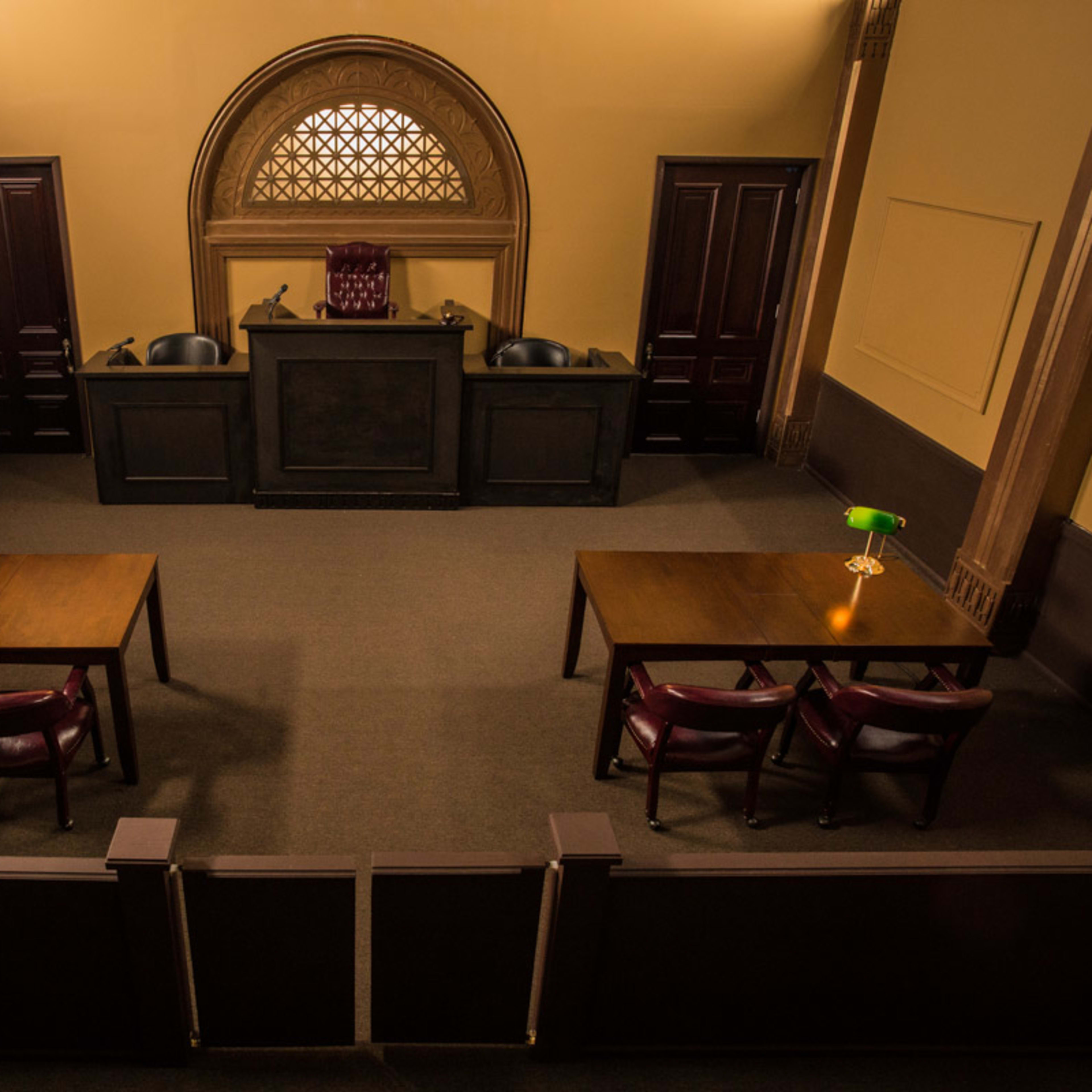 A courtroom interior with two wooden tables and chairs positioned in front of a judge's bench, lit by green desk lamps.