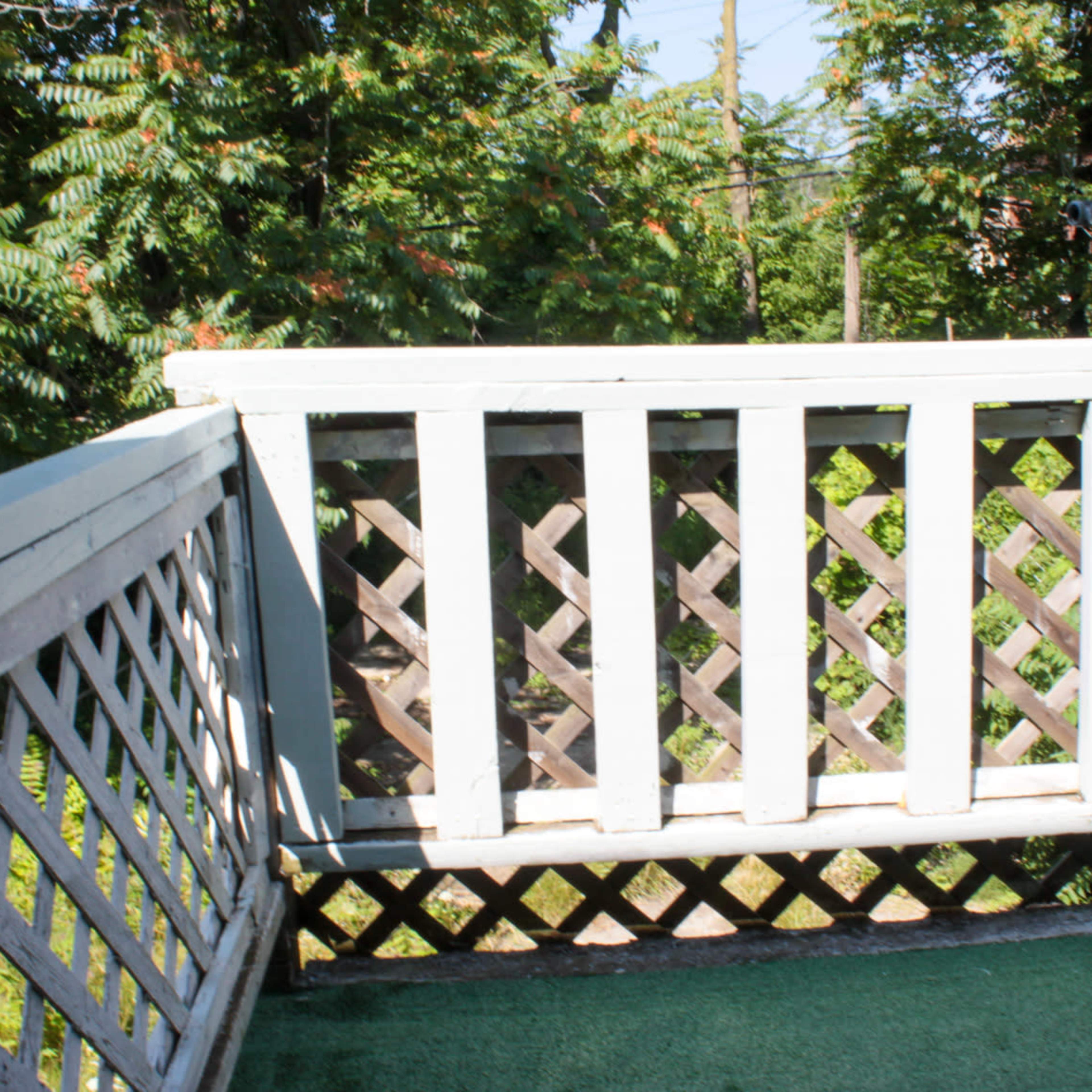 A wooden balcony with a white railing and a green carpet, overlooking greenery.