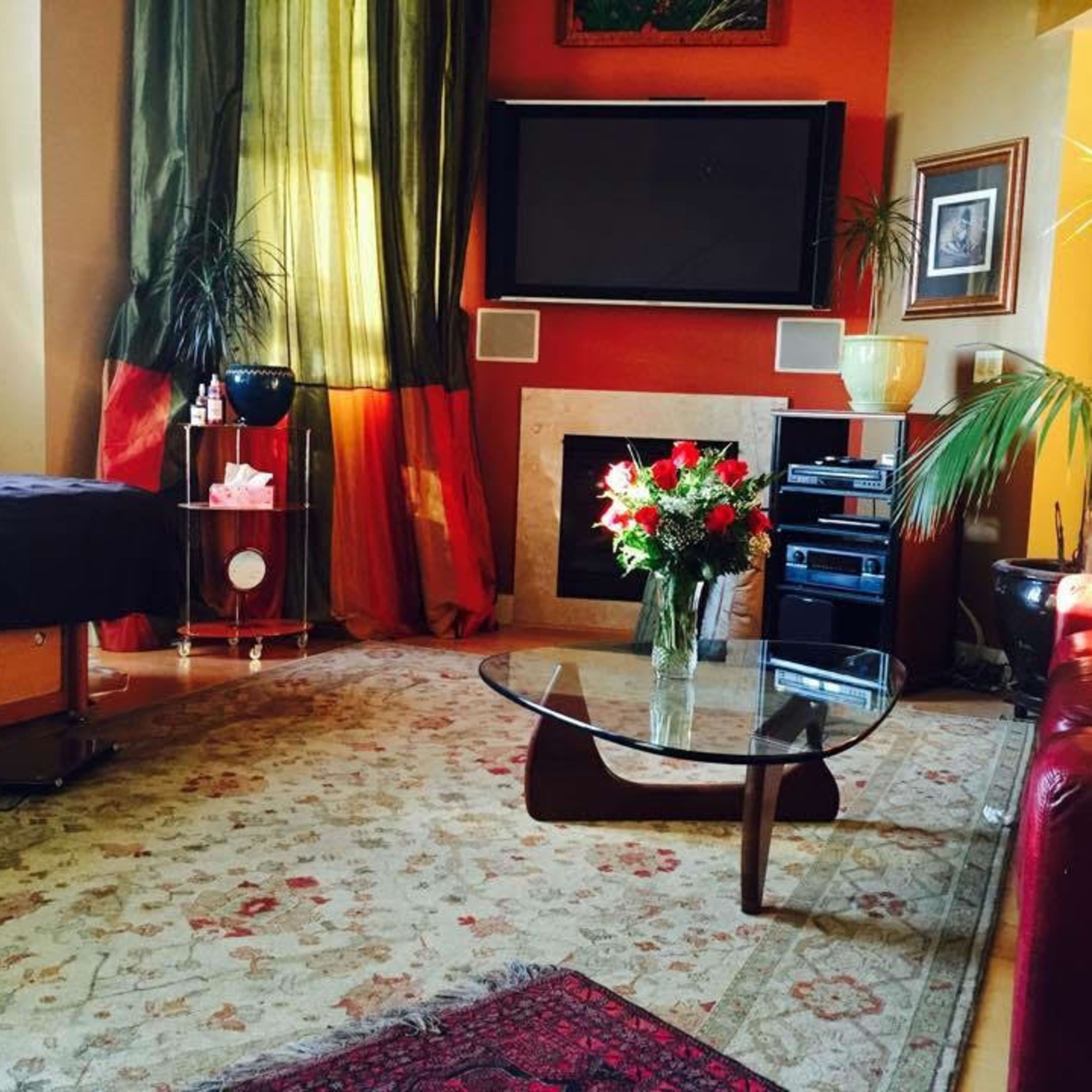A living room with a glass coffee table, a floral arrangement, and a television mounted on the wall, surrounded by decorative plants and warm-colored curtains.