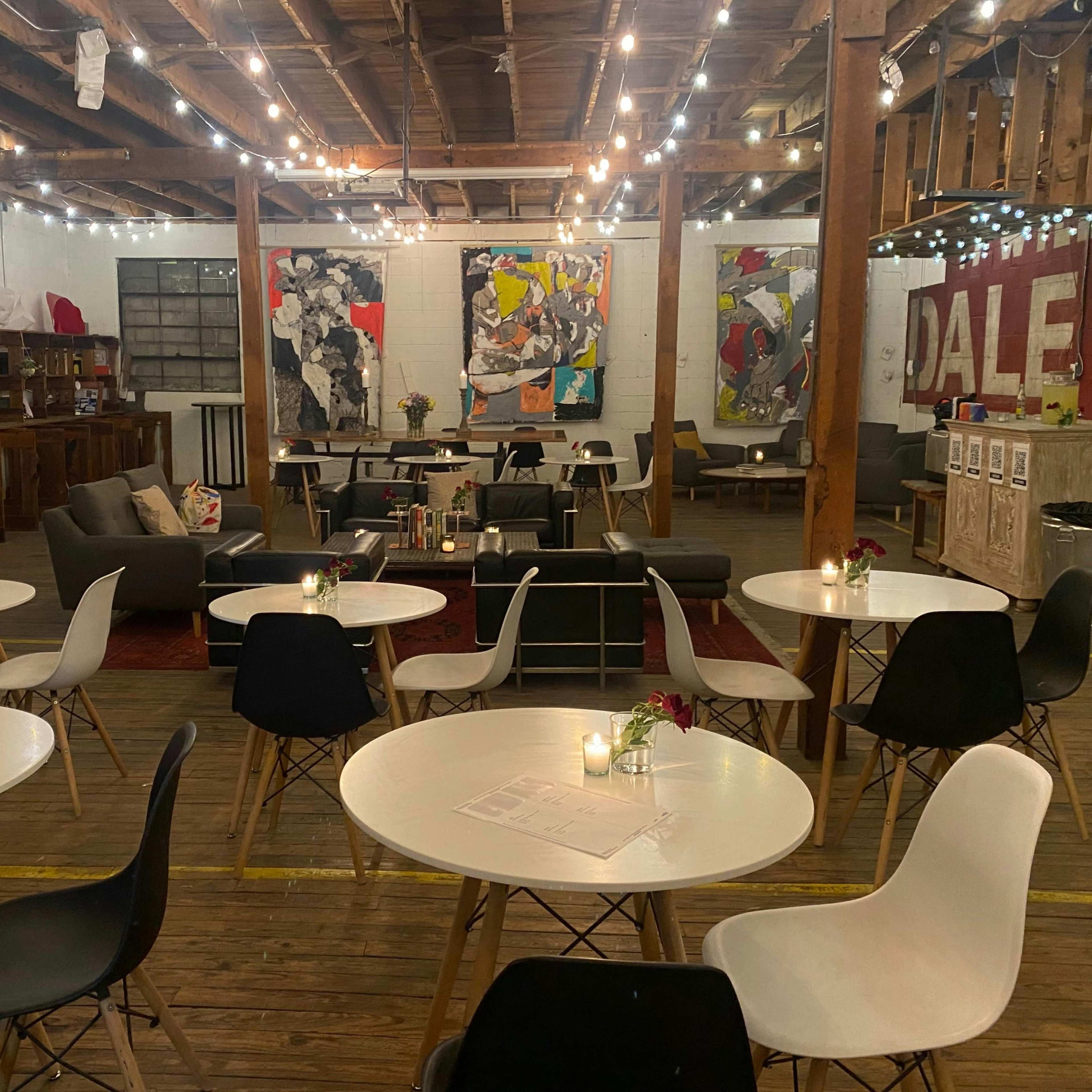 A spacious indoor venue with wooden floors, featuring tables with white tops and black and white chairs, illuminated by string lights and adorned with artwork on the walls.