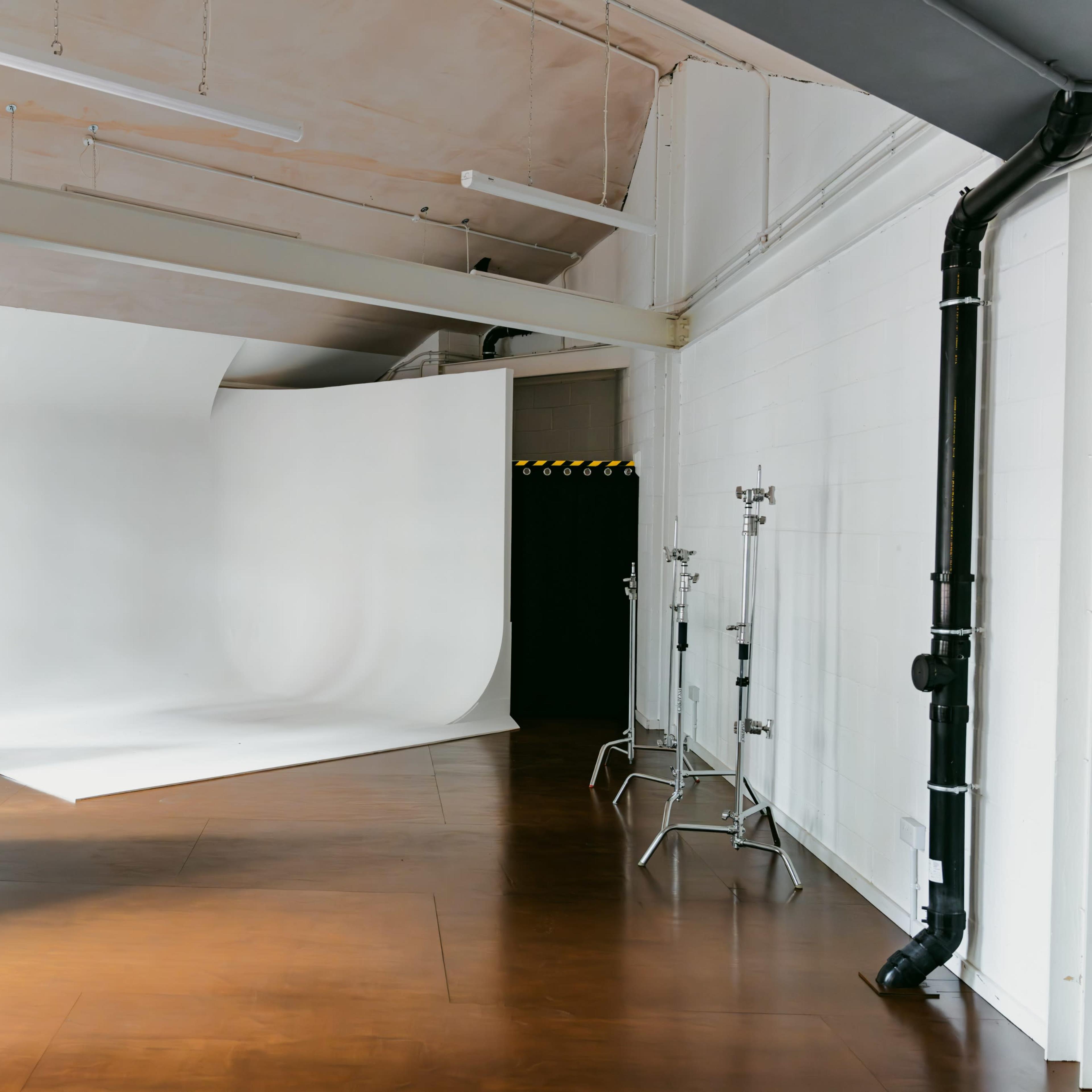A spacious studio with a white backdrop, wooden flooring, and studio equipment in the corner.