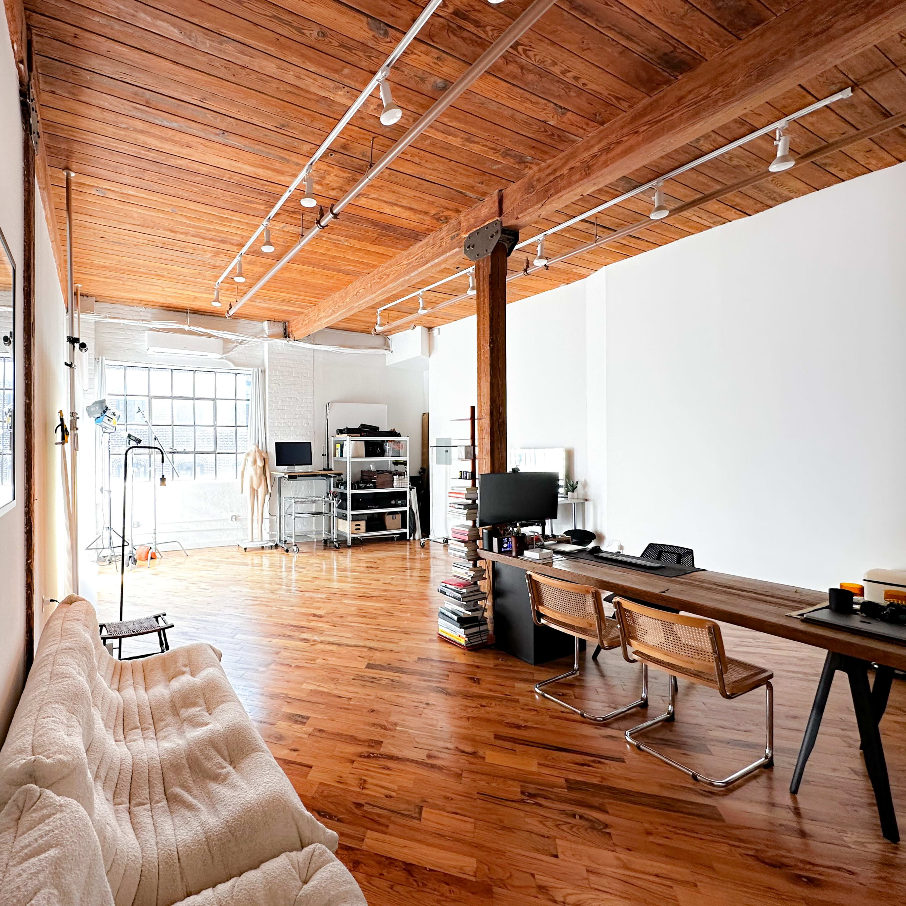 A spacious room with wooden floors, large windows, and minimal furniture, including a desk with chairs and a sofa.