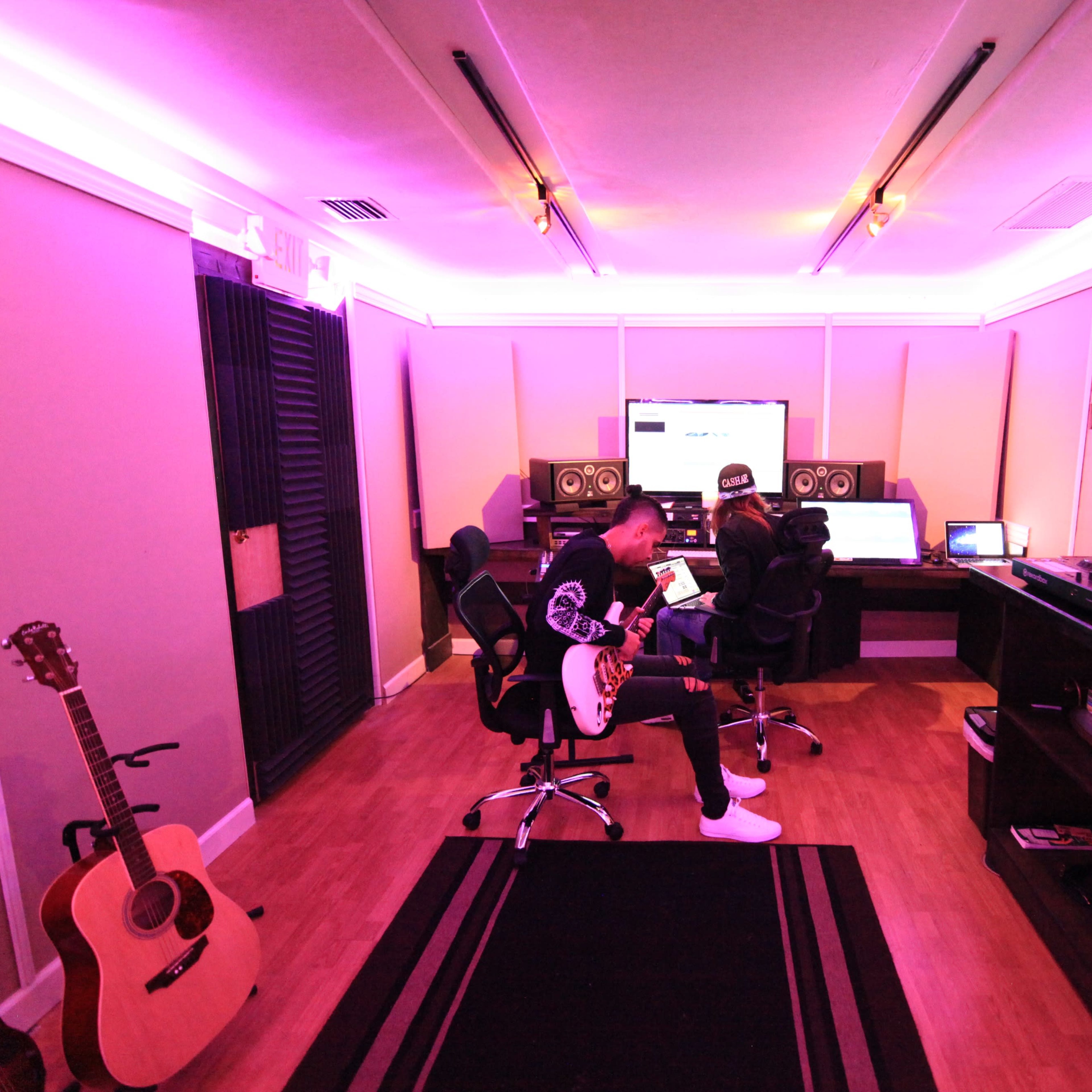 A musician plays guitar in a music studio with computers, speakers, and lighting enhanced by purple accents.