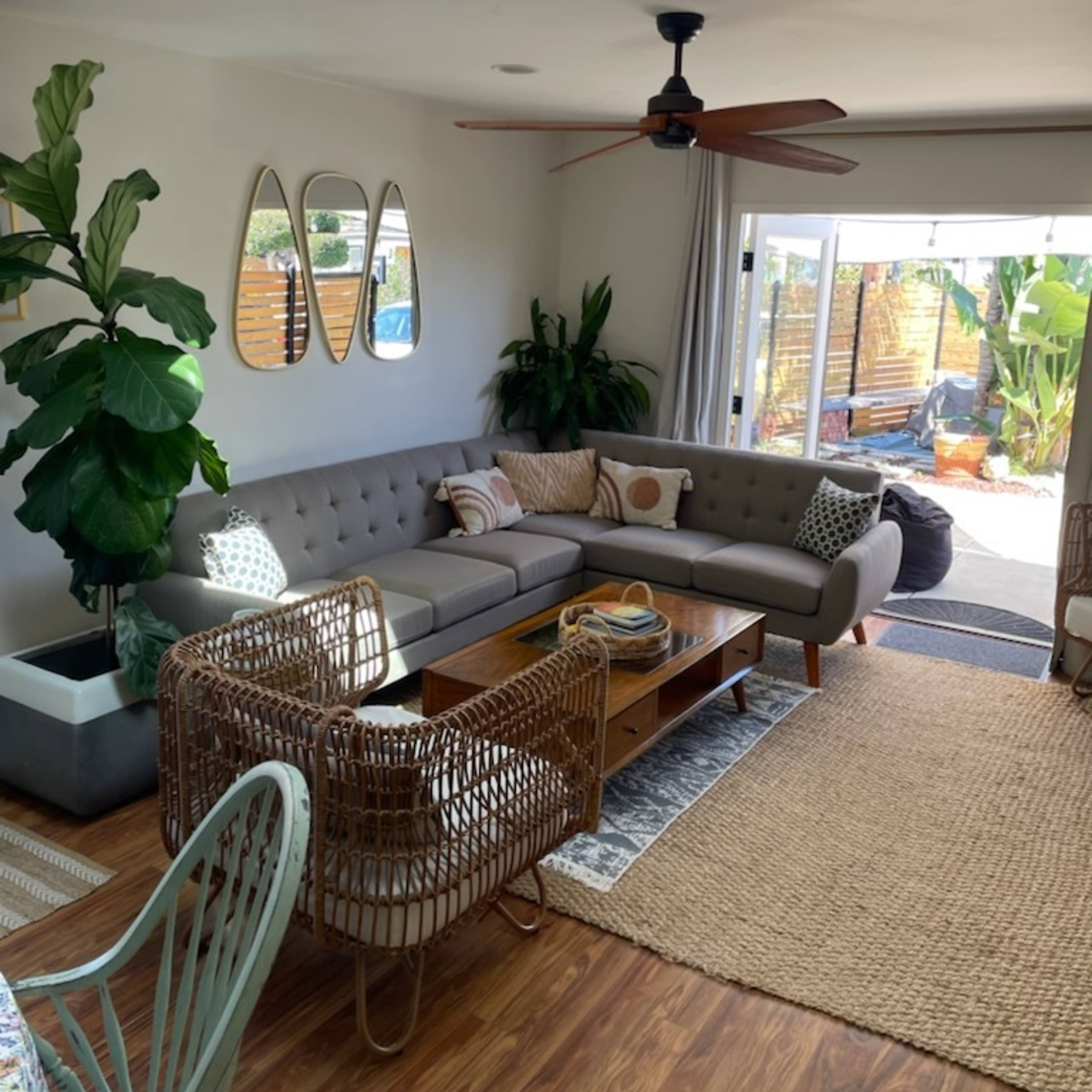 The living room features a sectional sofa, two woven chairs, a coffee table, and large windows opening to a patio with plants.