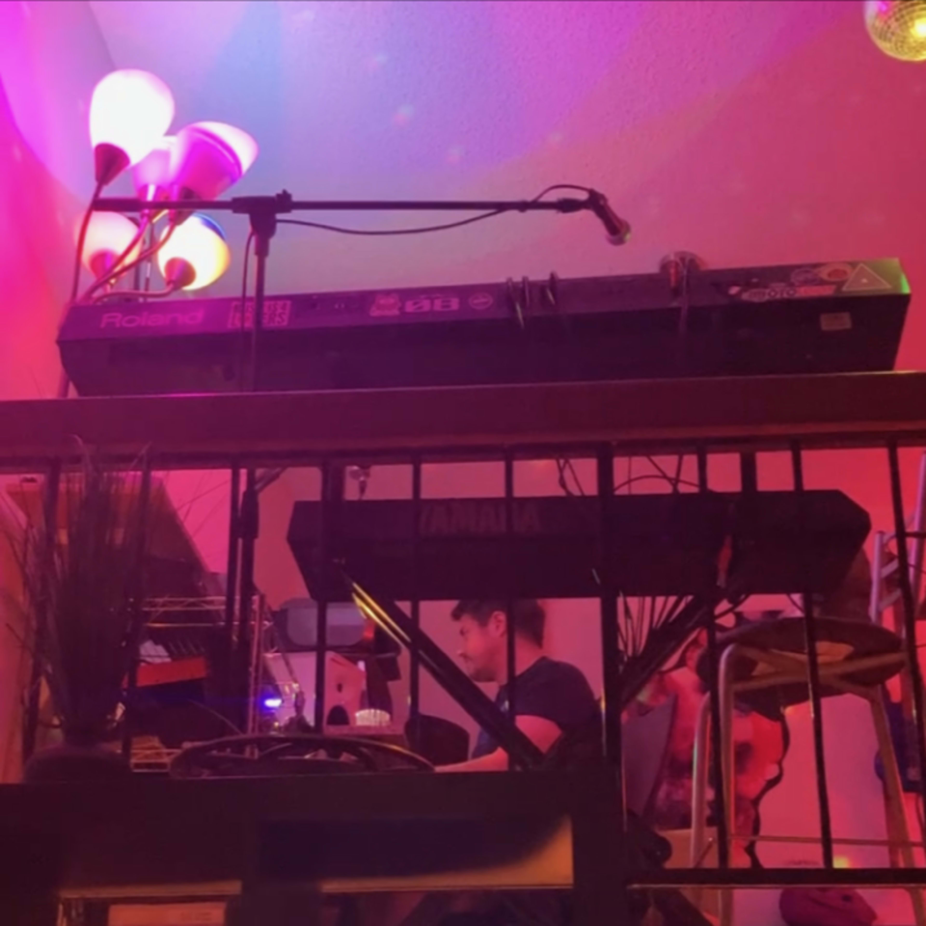 A musician sits at a keyboard setup on a balcony, with colorful lights illuminating the scene and pixel art characters visible below.