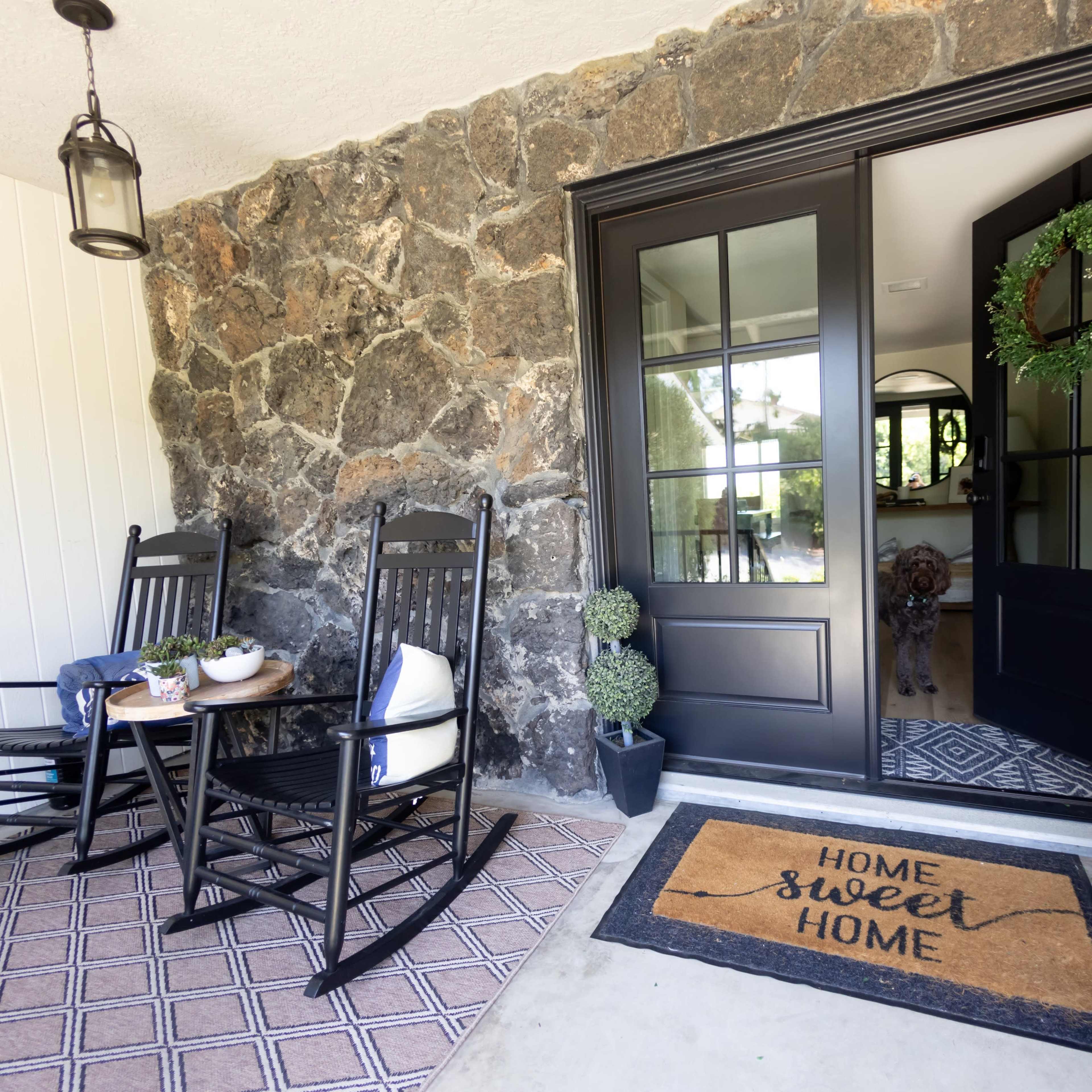 A stone-walled porch features two rocking chairs, a decorative welcome mat, and a view of a dog standing inside the house.