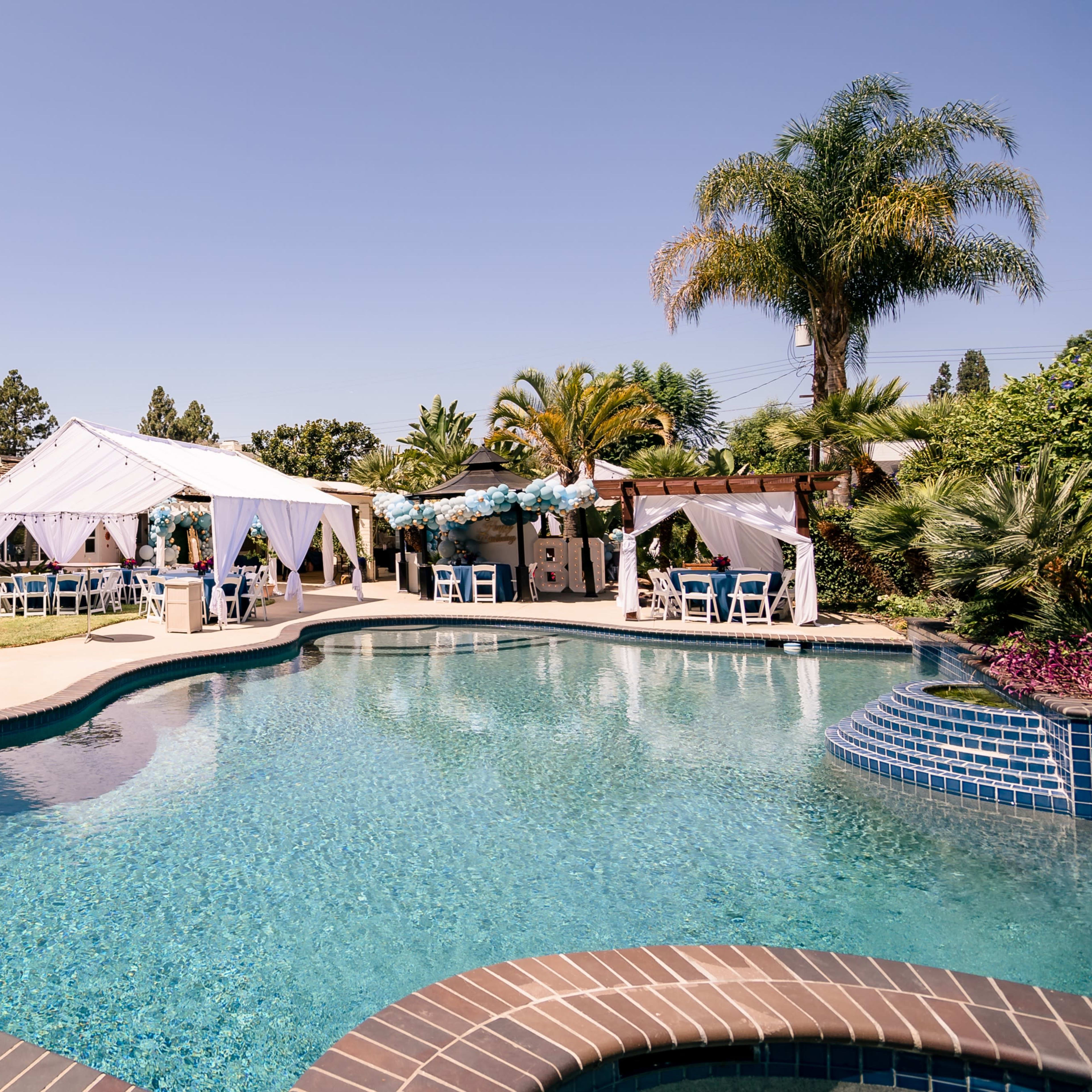 A clear swimming pool is surrounded by tents and seating in a landscaped backyard.