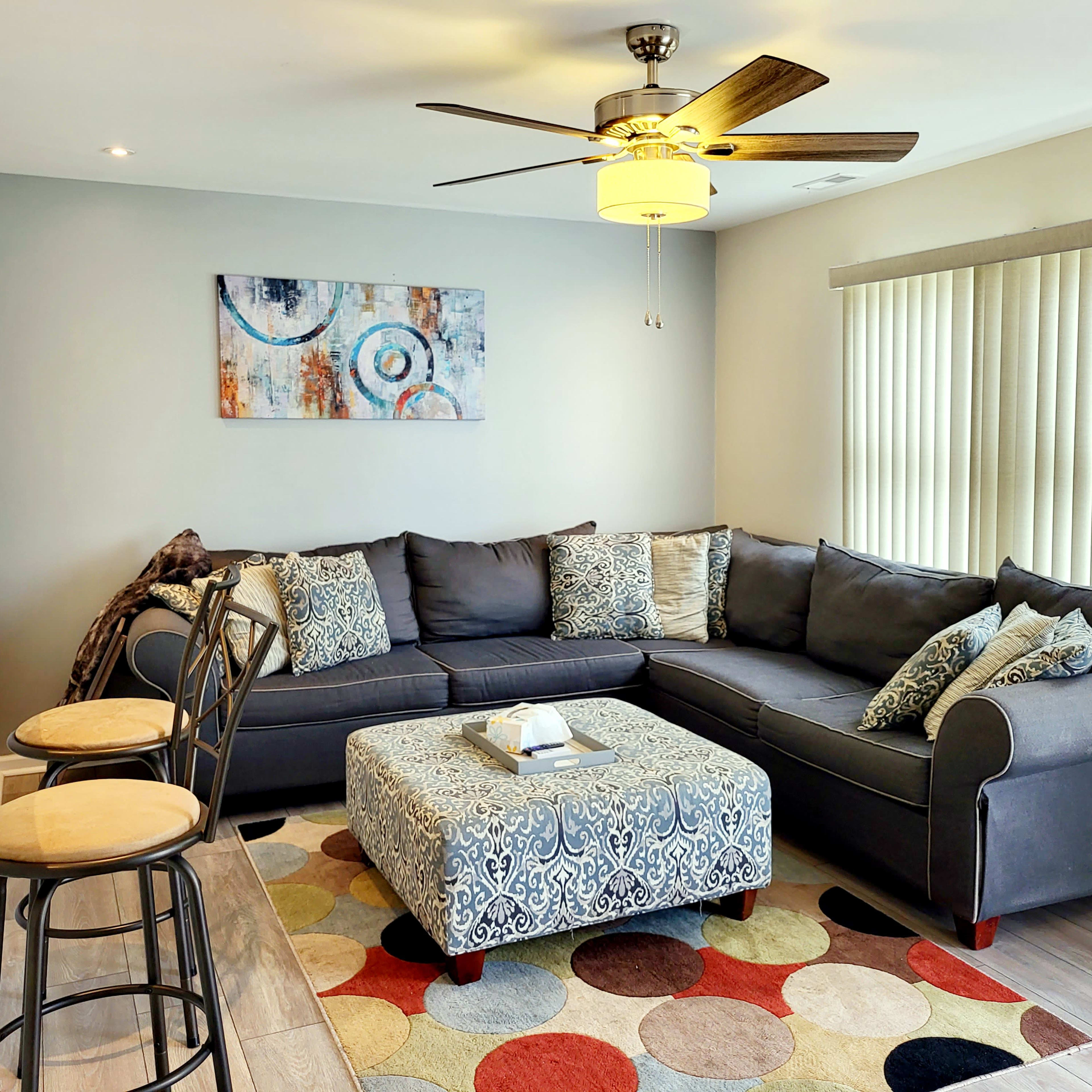 The living room features a gray sectional sofa with decorative pillows, an ottoman, a circular rug, and a ceiling fan, arranged around a bright window with vertical blinds.