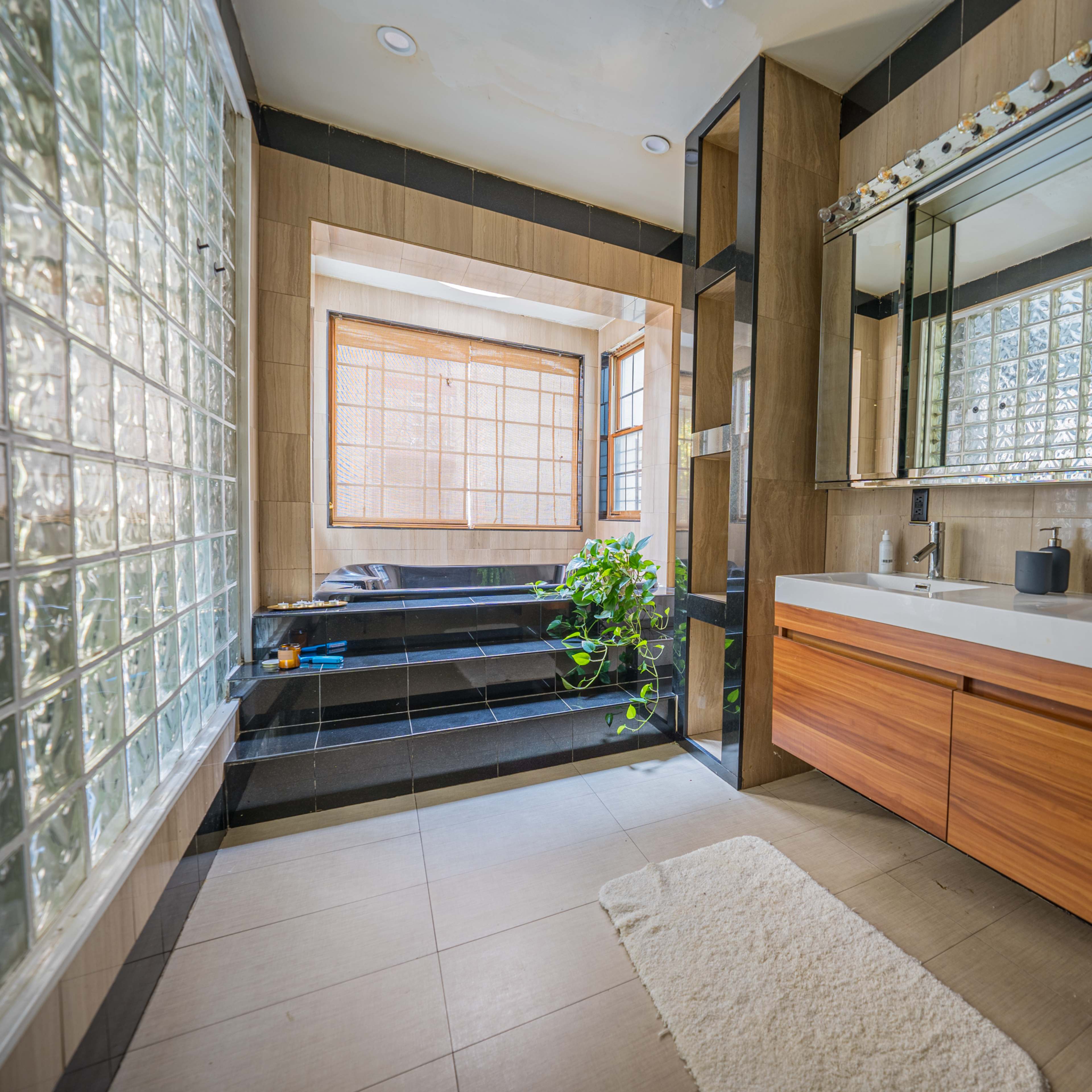 A modern bathroom featuring a double sink vanity with wooden cabinets, a glass block window, and a view of a small indoor seating area with plants.
