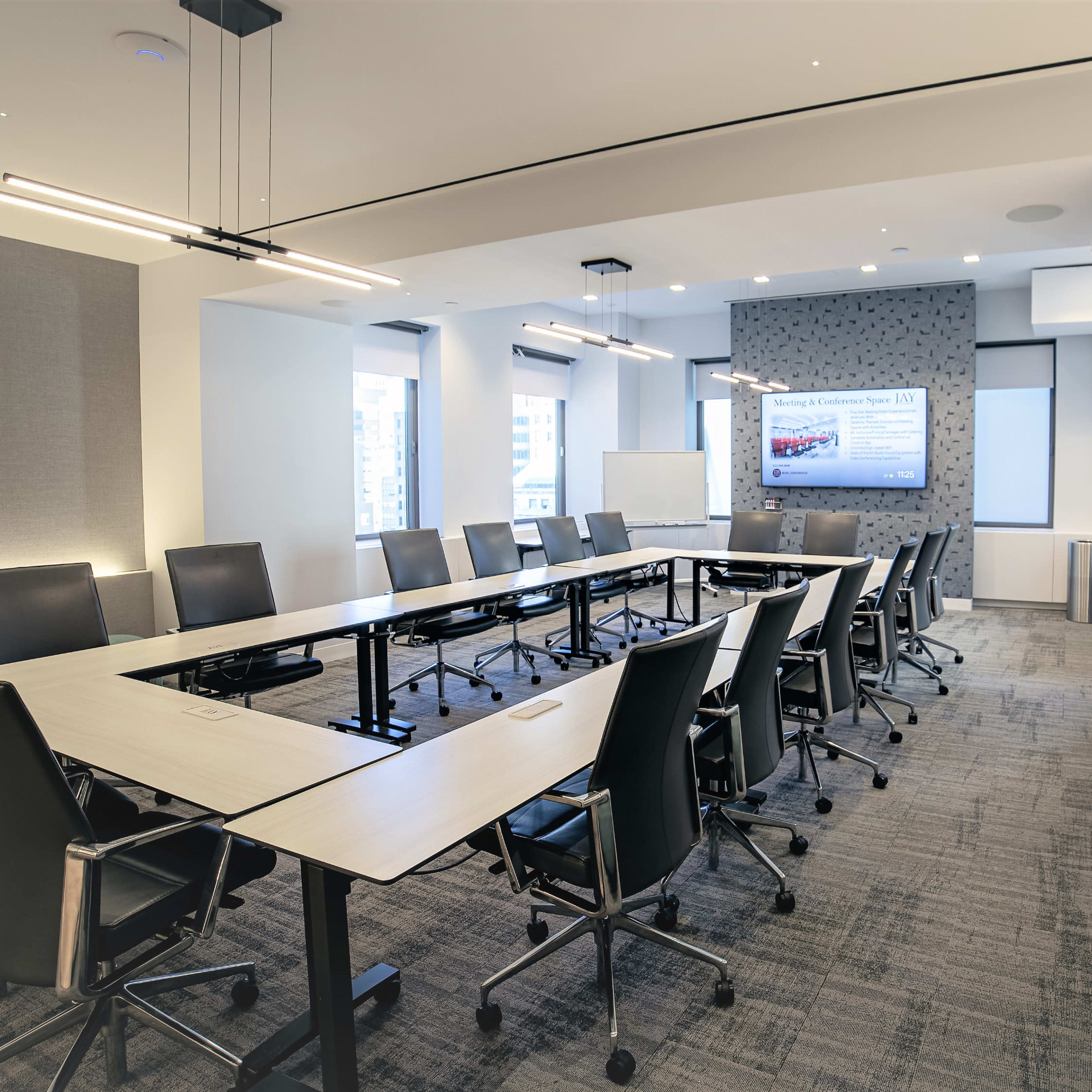 A modern conference room features a long table surrounded by black chairs, with large windows and a wall-mounted screen displaying a presentation.