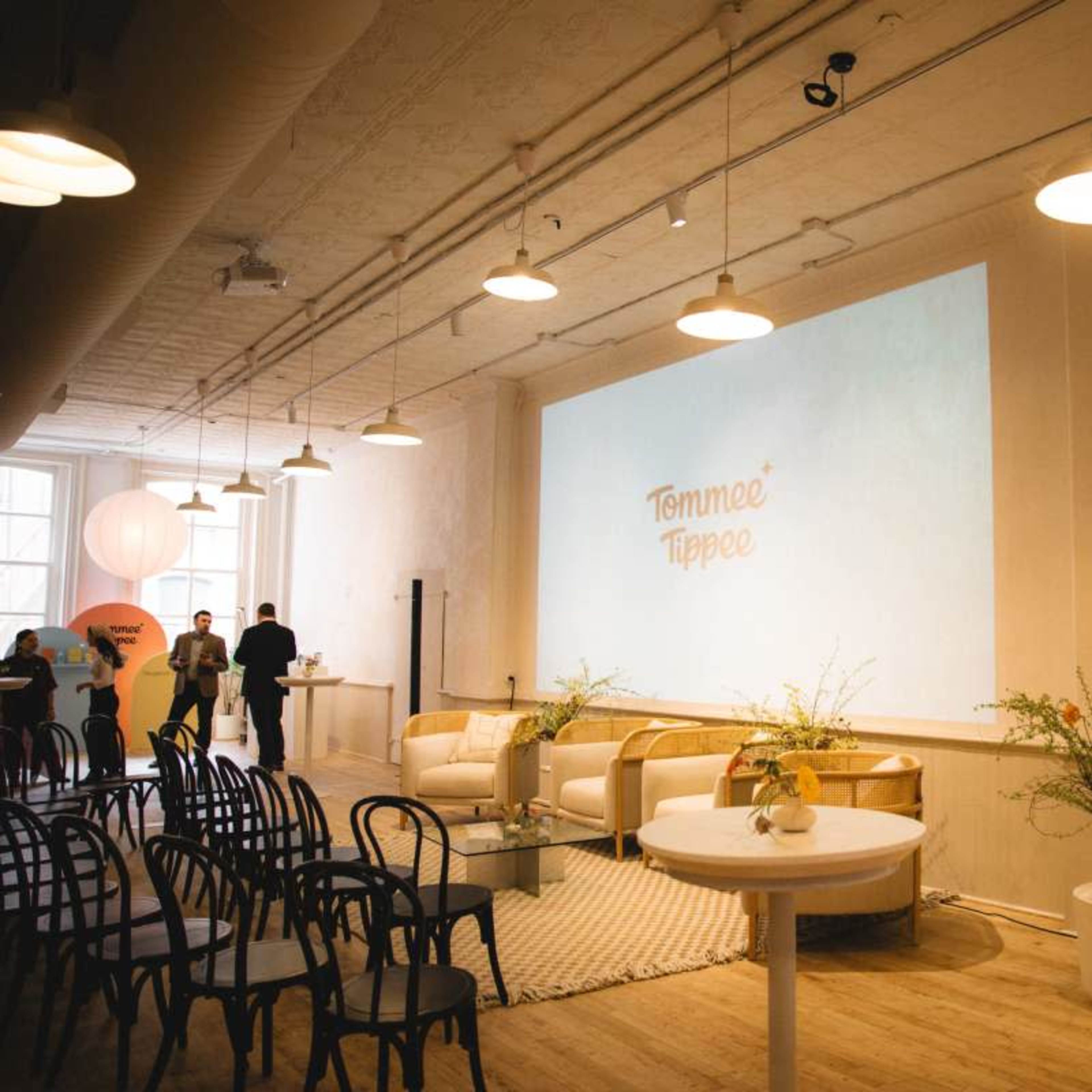 A well-lit event space set up with chairs, a large screen displaying a logo, and various seating arrangements.