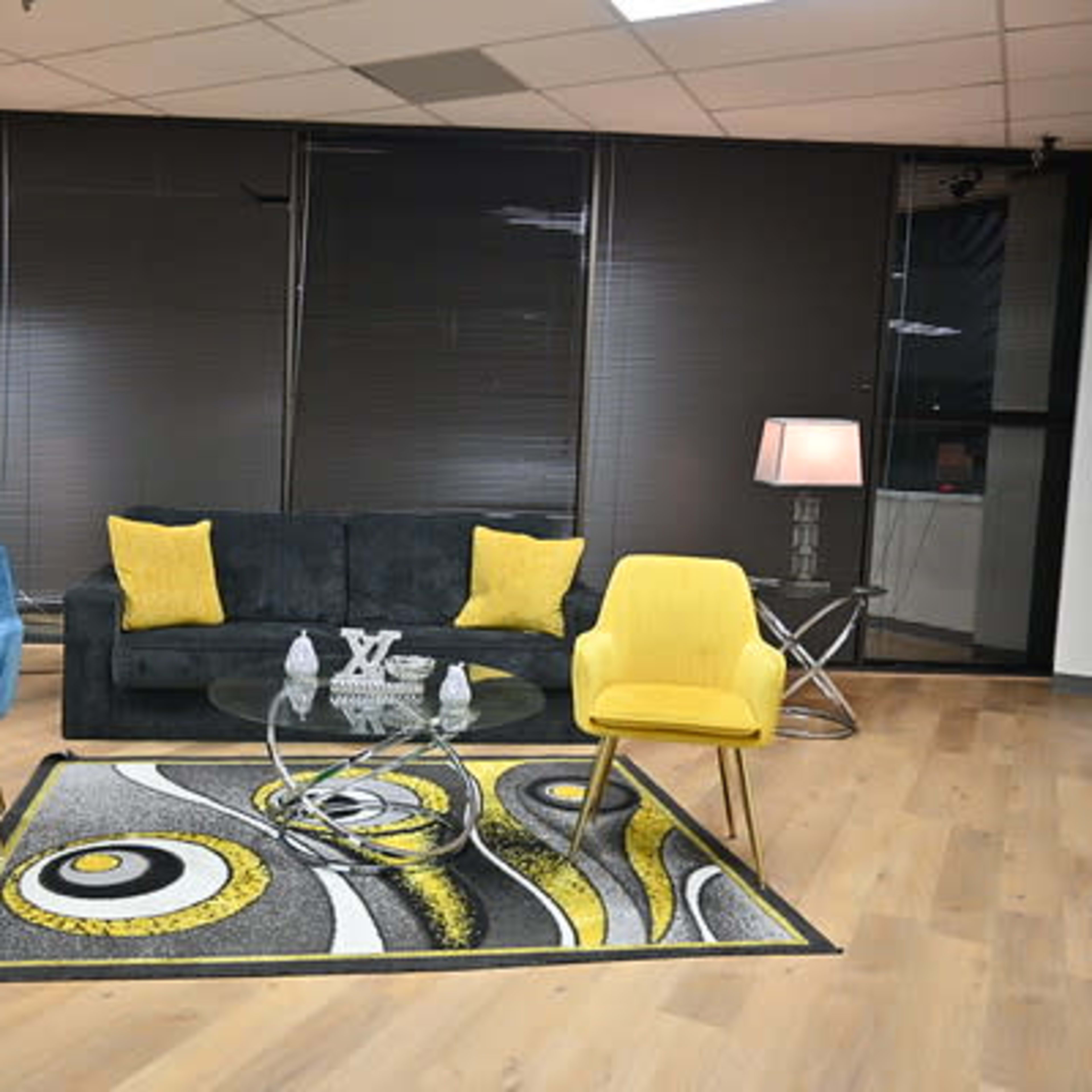A modern living room setup featuring a black sofa, two colorful chairs, a round glass coffee table, and a patterned rug.