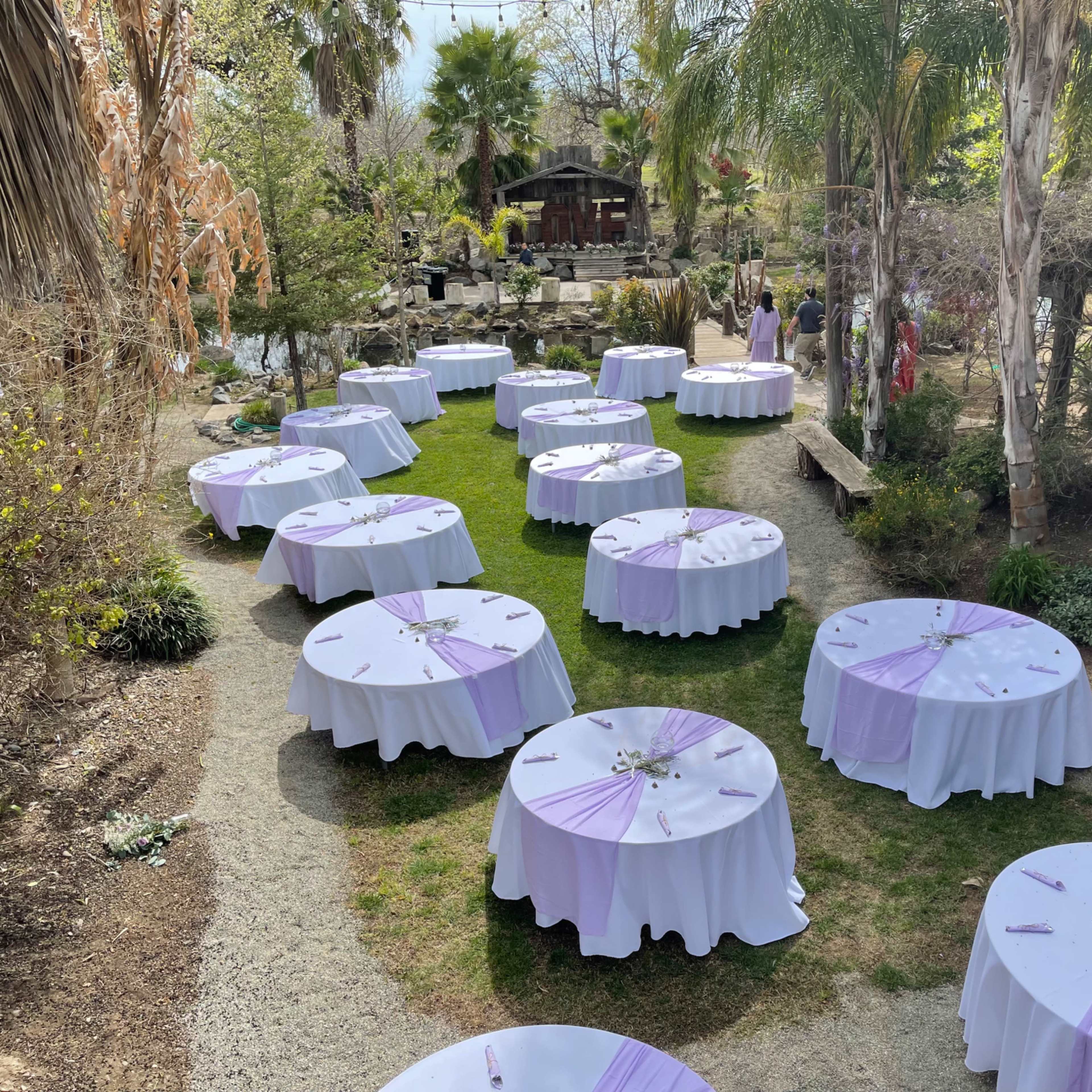 A garden venue set up for an outdoor event with round tables covered in white tablecloths and lavender accents arranged on a grassy area.