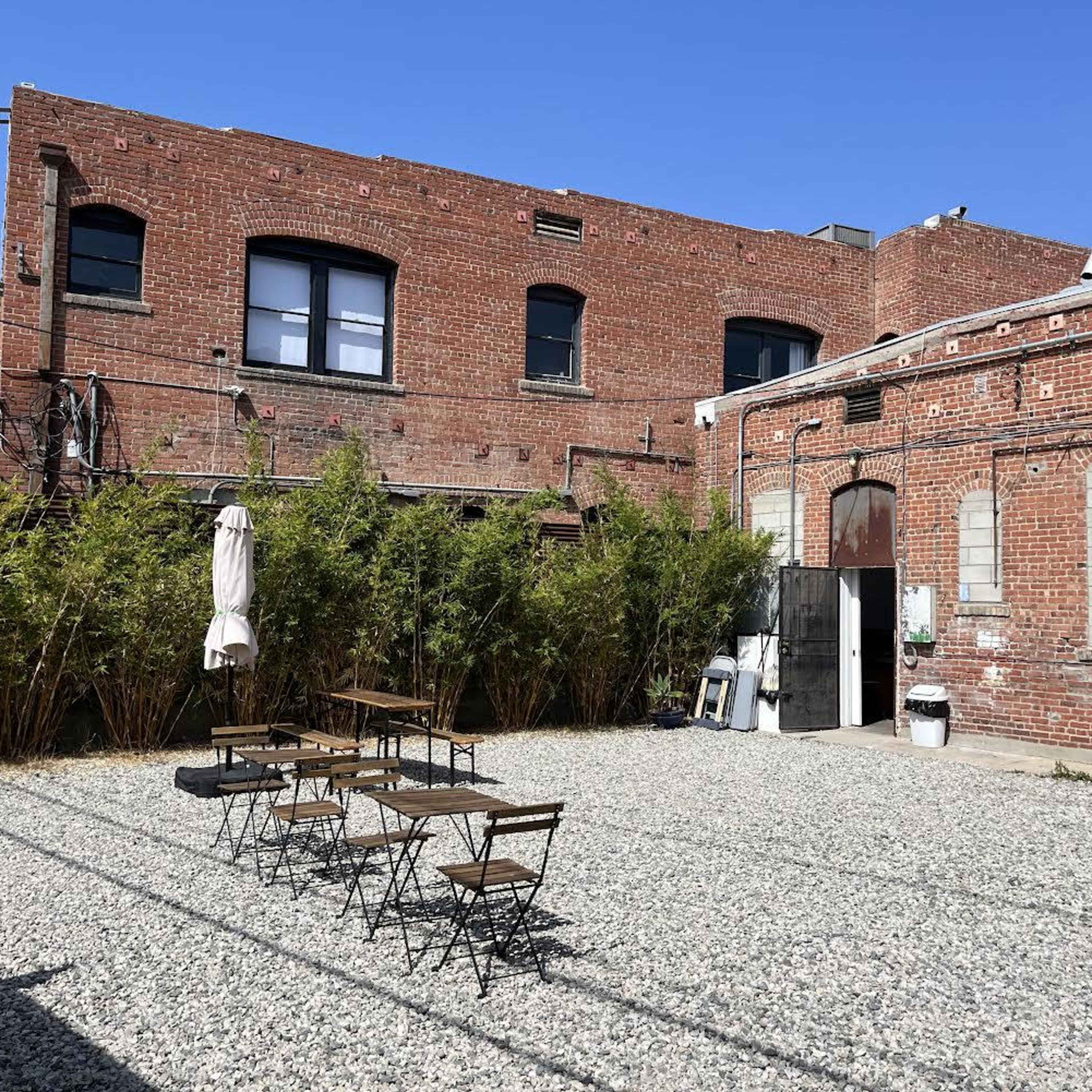 A gravel courtyard features several metal tables and chairs surrounded by a brick building and bamboo plants.