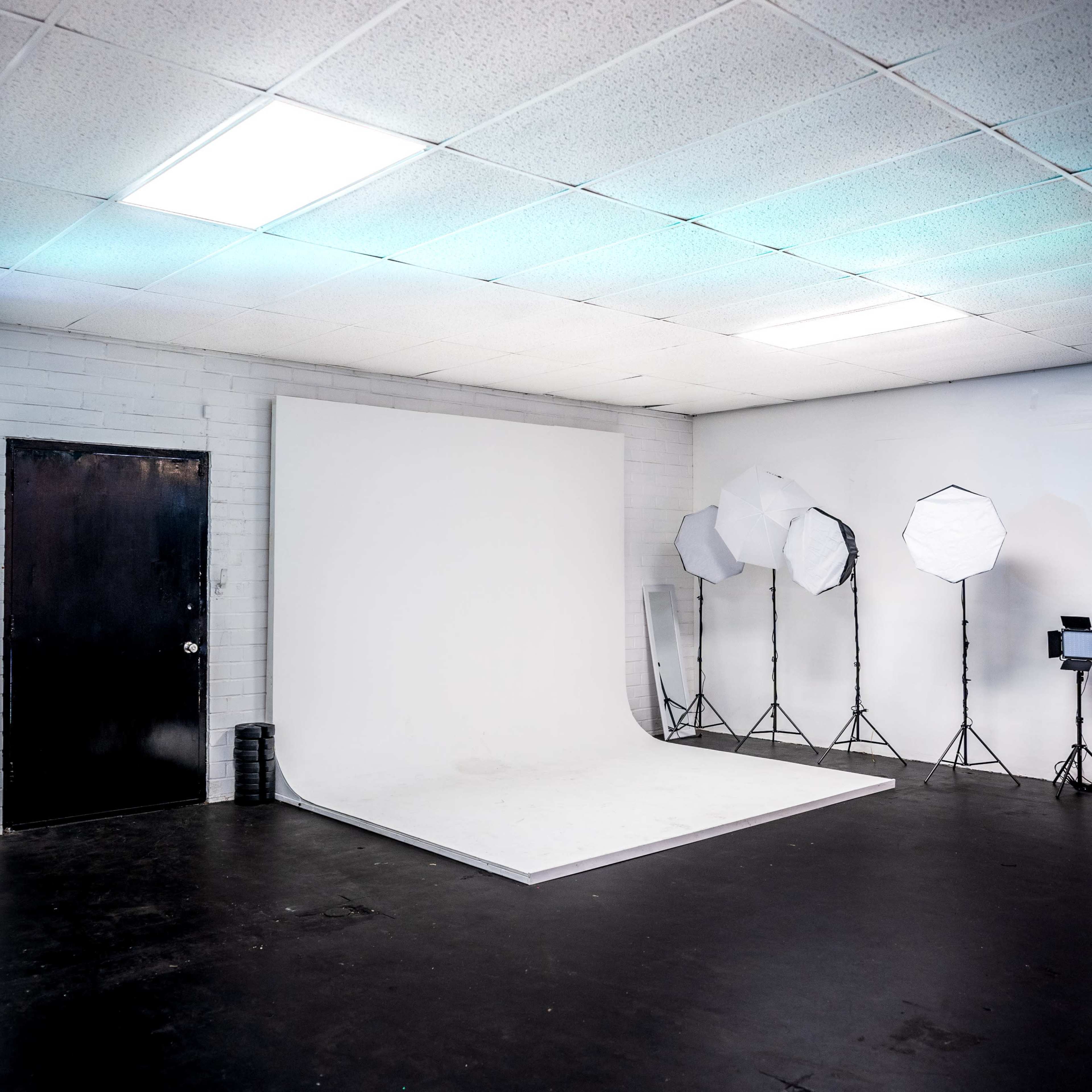 A photo studio featuring a white backdrop, lighting equipment on tripods, and a dark door in a room with a black floor and ceiling tiles.