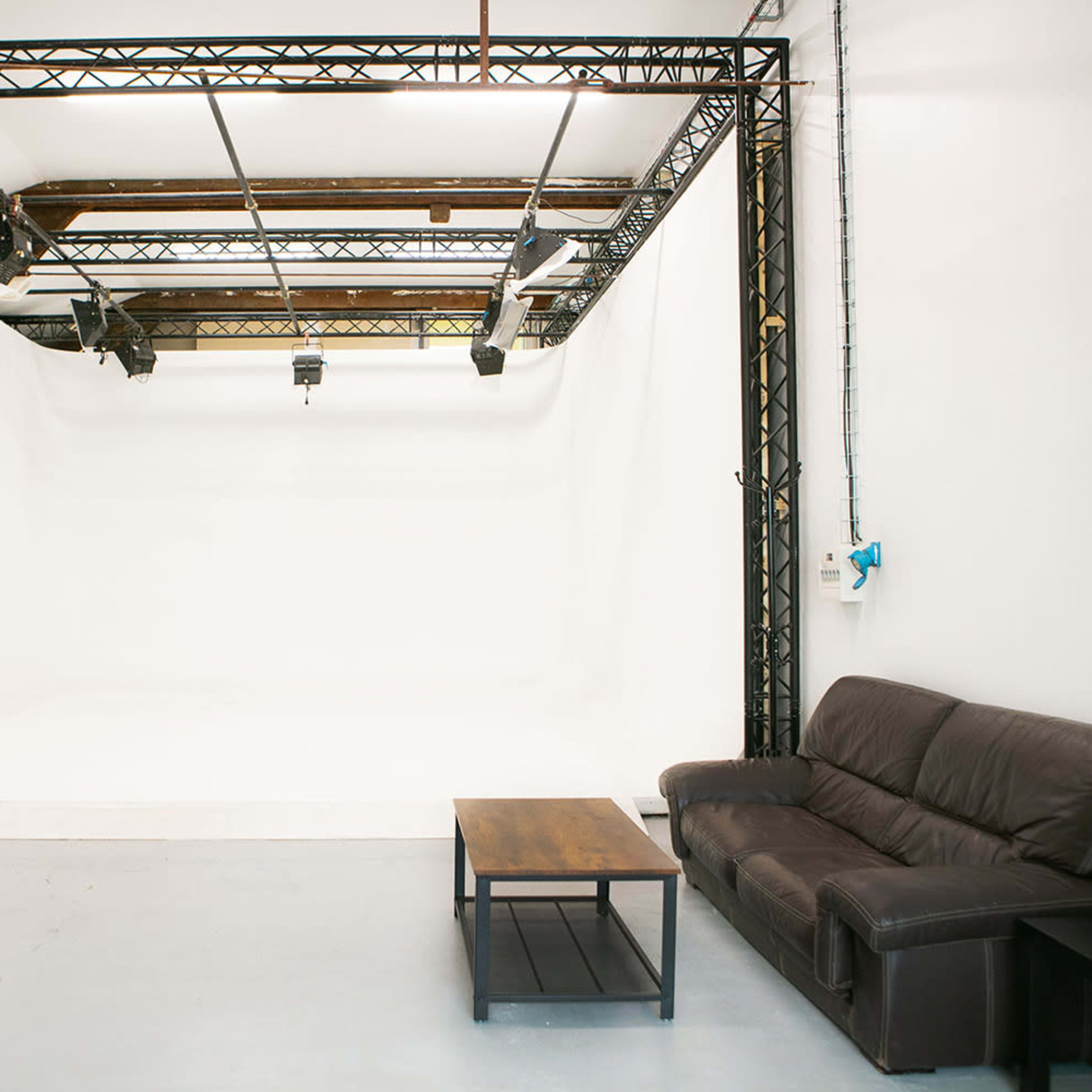 A photography studio with a large white backdrop, black support beams, and a brown leather couch next to a wooden coffee table.