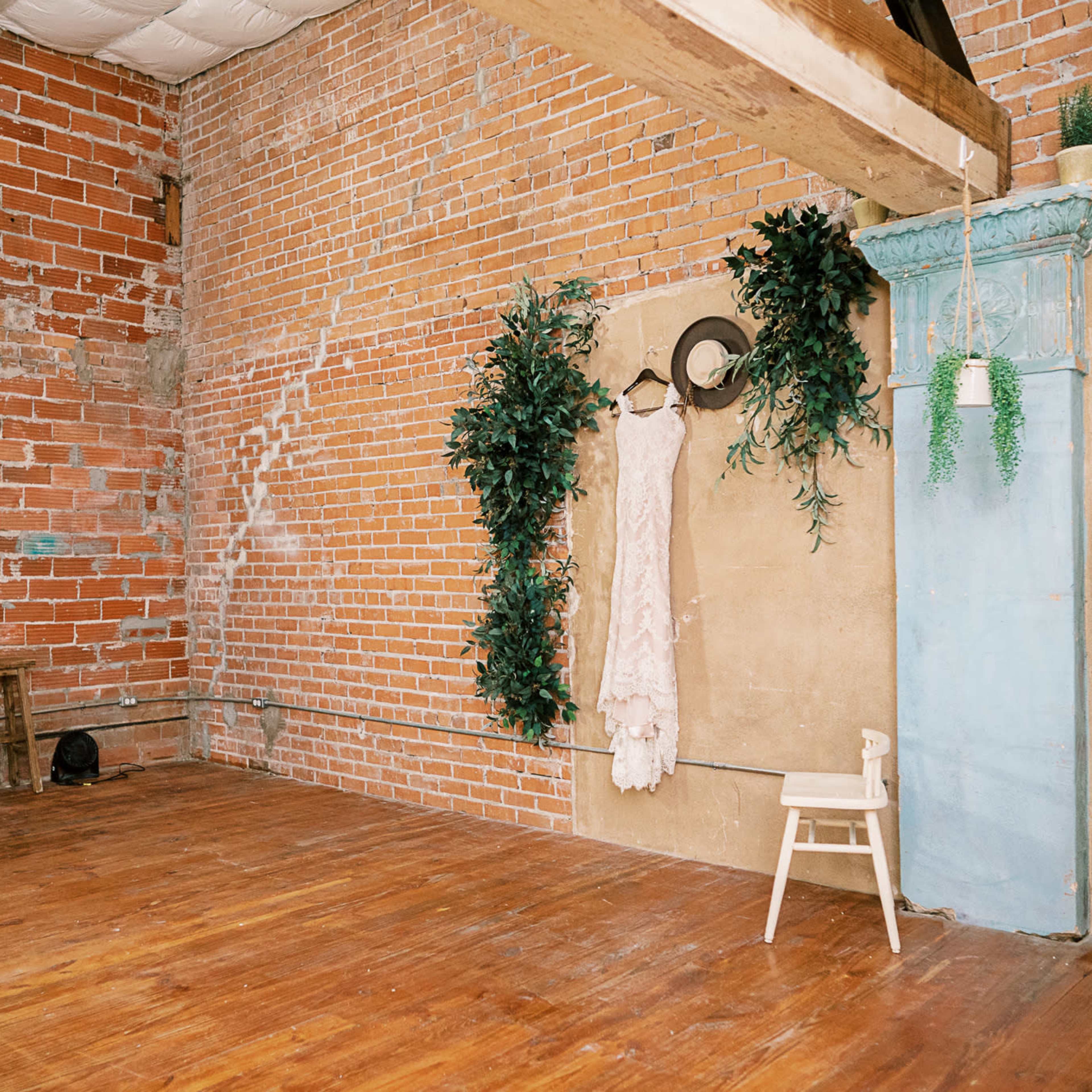 A spacious, unfurnished room with exposed brick walls, a wooden floor, a small table with chairs, and a hanging dress next to a blue decorative column.