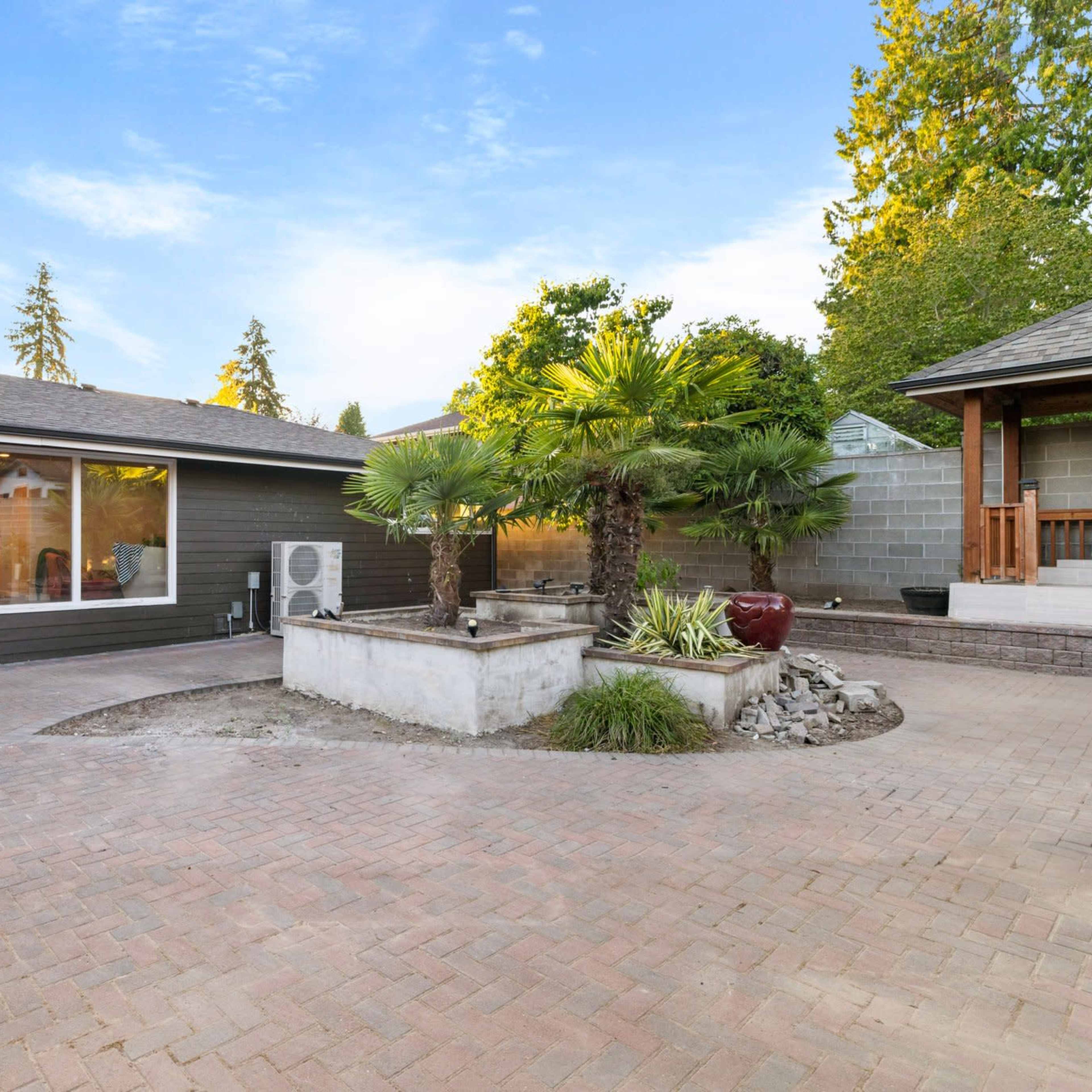 A spacious outdoor patio area featuring a brick surface, a small fountain with plants, and a neighboring structure with a shaded porch.