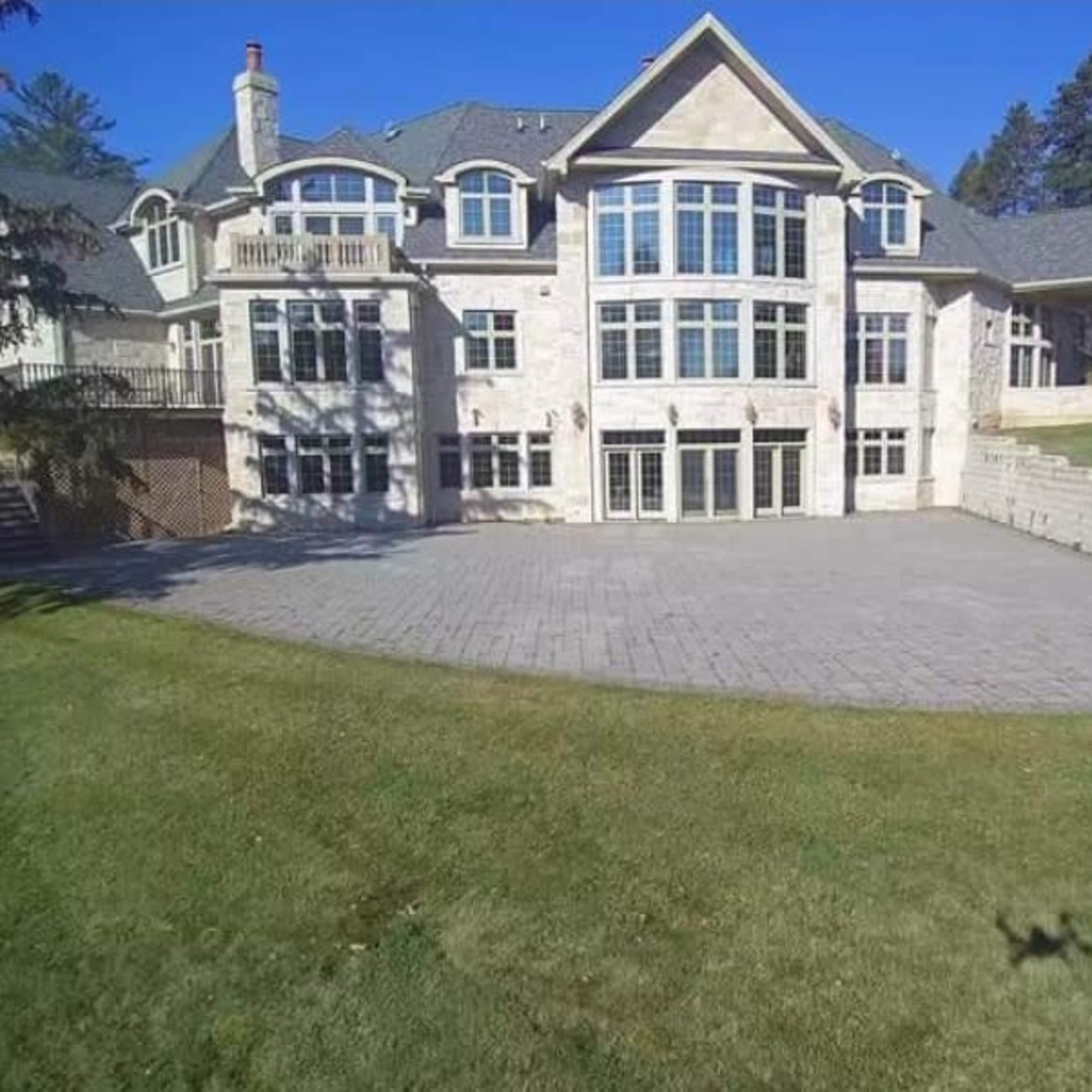 A large, multi-story stone house with numerous windows sits on a grassy lawn, featuring a spacious patio area in the foreground.