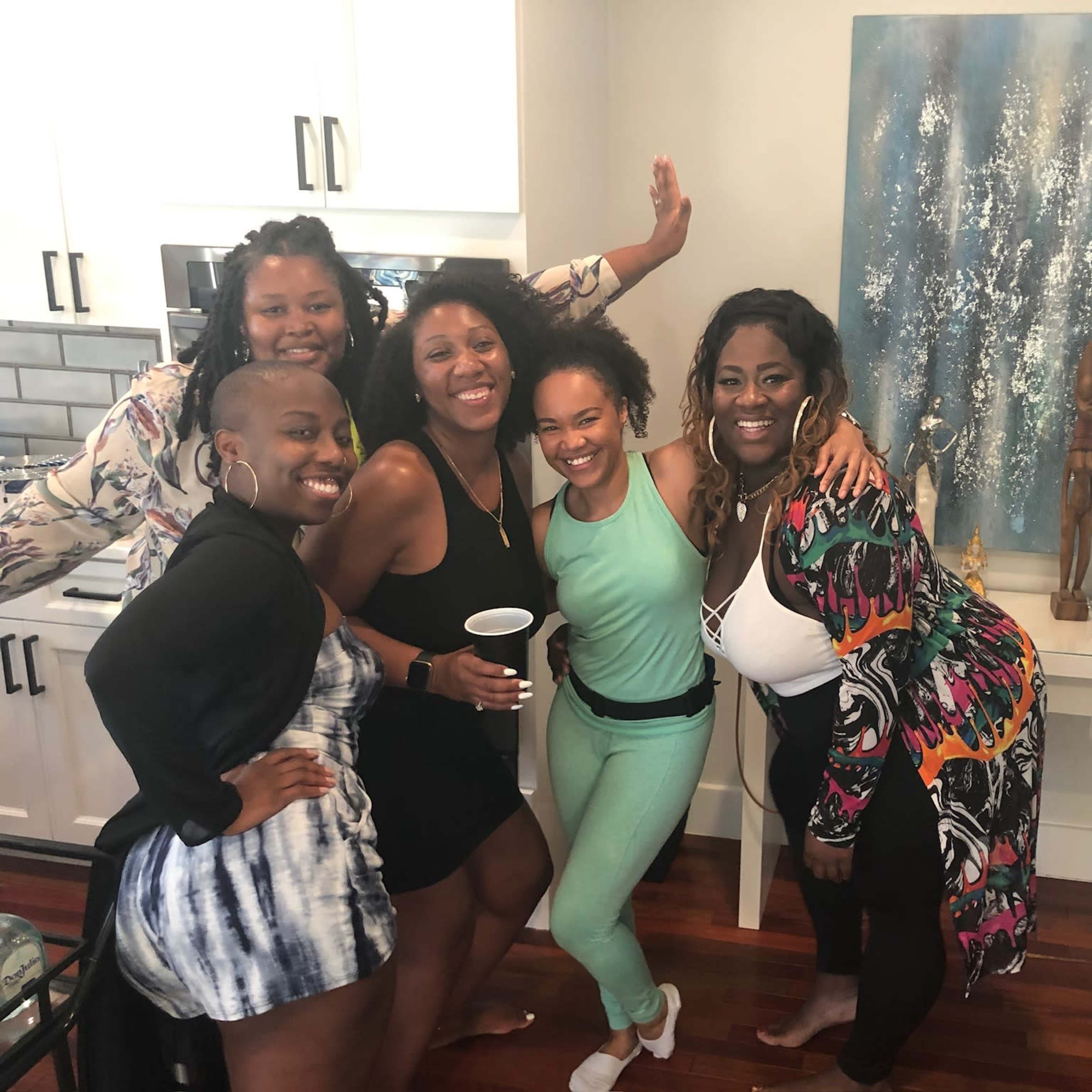 Five women gather in a kitchen, smiling and posing together in front of a light blue painting on the wall.