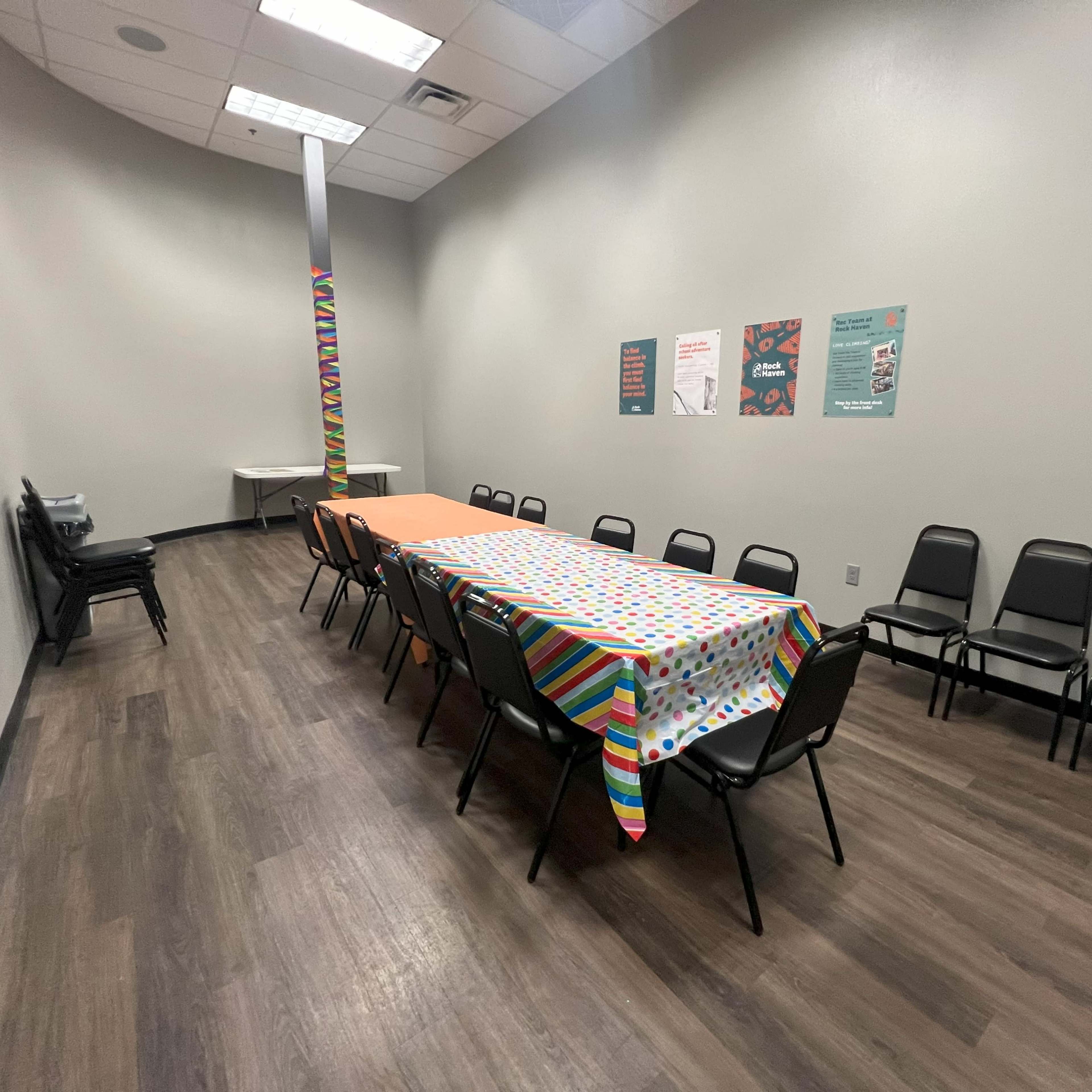 A long table covered with a patterned tablecloth is set up in a room with gray walls and black chairs arranged around it.