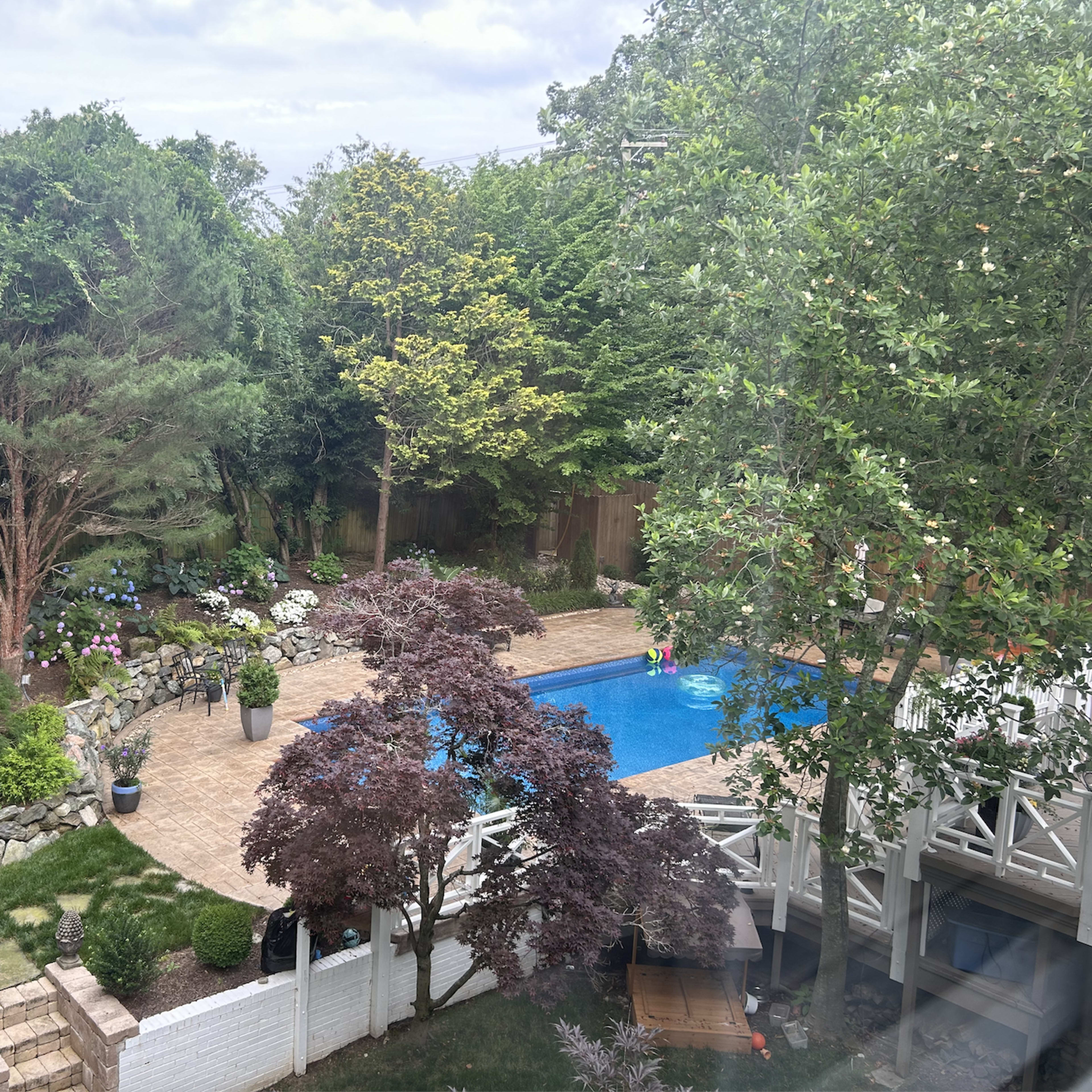 A backyard features a swimming pool surrounded by trees, flowering plants, and a patio area.