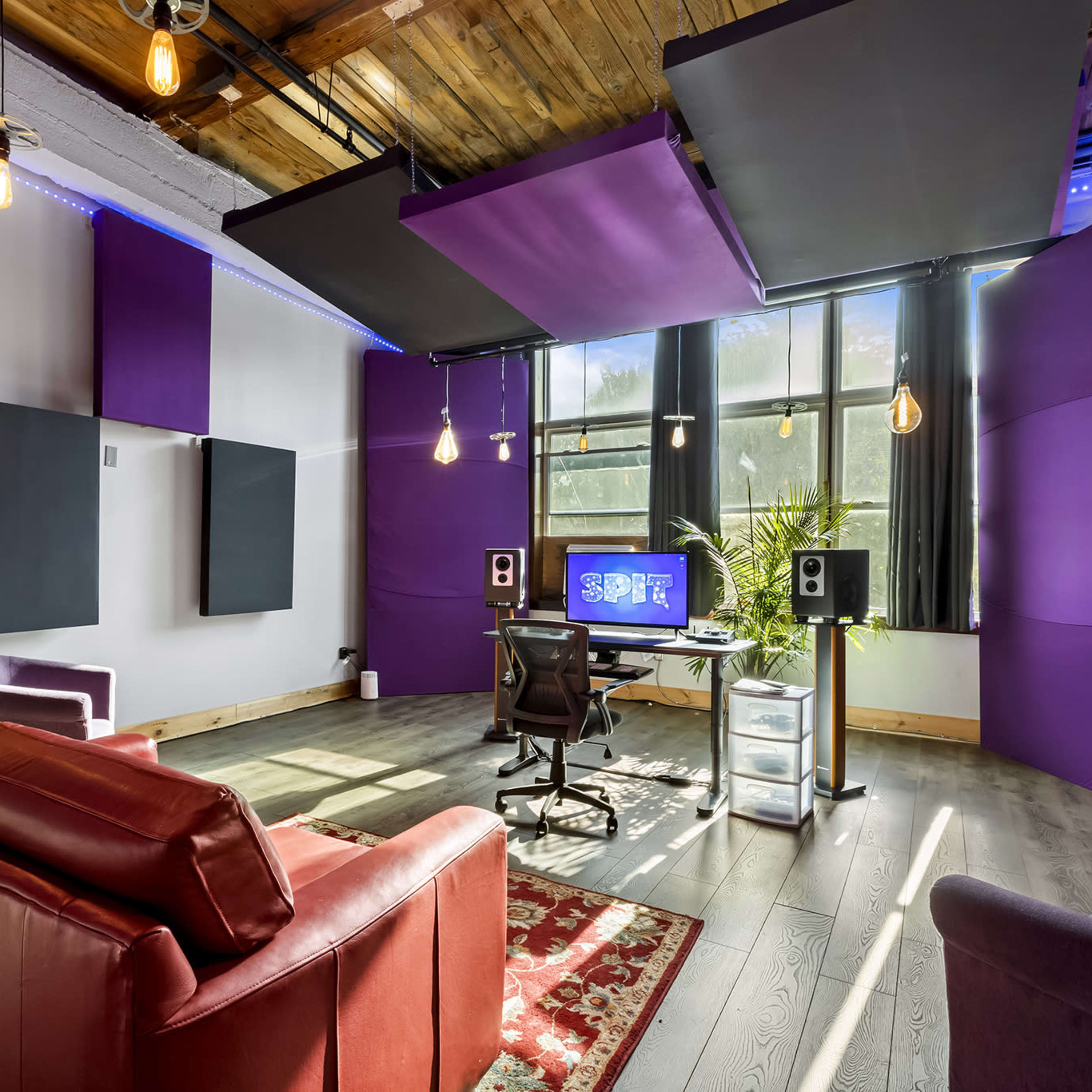 A modern studio space with purple acoustic wall panels, a desk with a computer setup, and a seating area featuring a red couch and a decorative rug.
