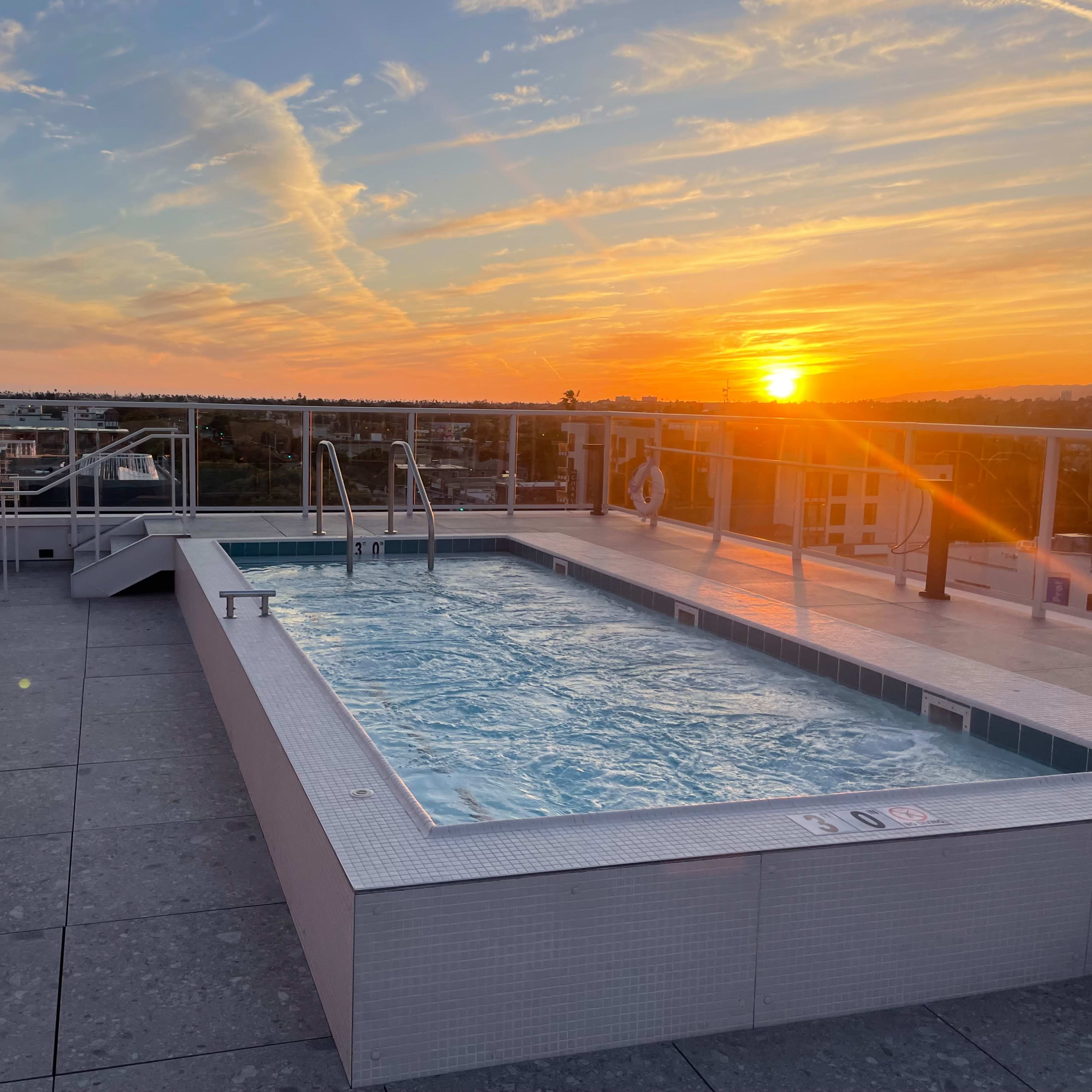 A rooftop pool with clear water reflects the vibrant colors of a sunset over a city skyline.