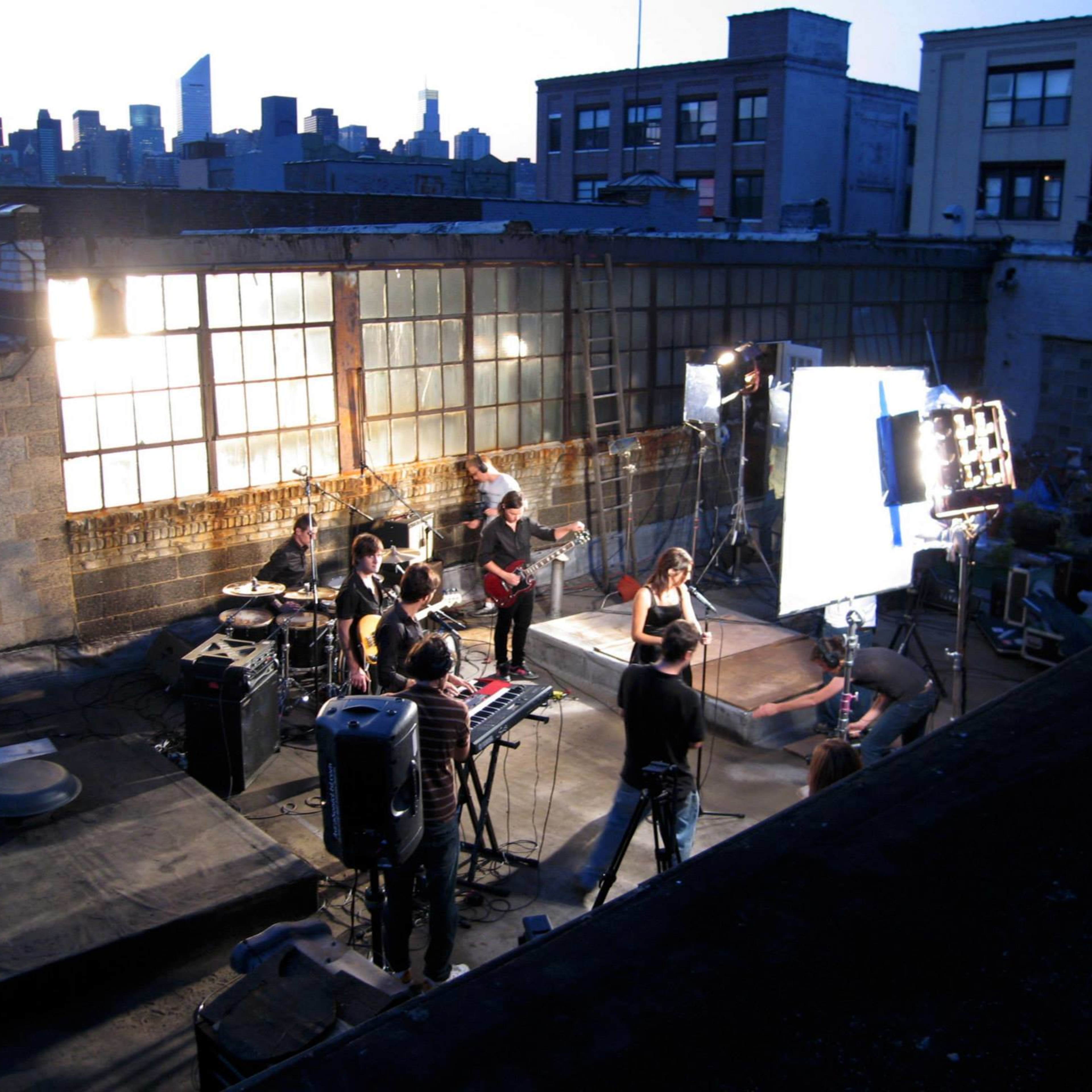 A group of musicians and crew members are setting up for a performance on a rooftop, with city buildings visible in the background.
