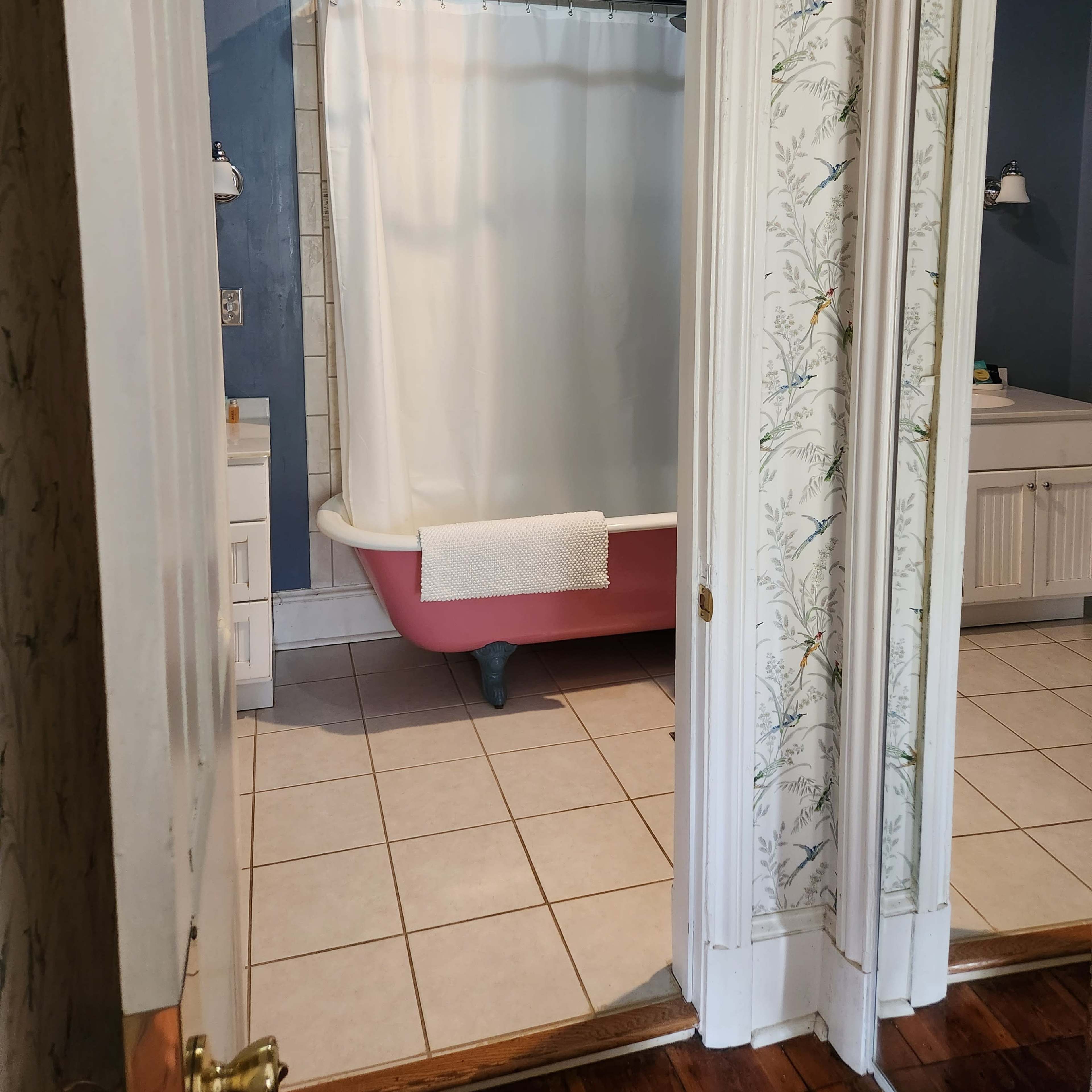 A pink bathtub with a white shower curtain is positioned in a bathroom, visible through a door framed by white woodwork and surrounded by patterned wallpaper.