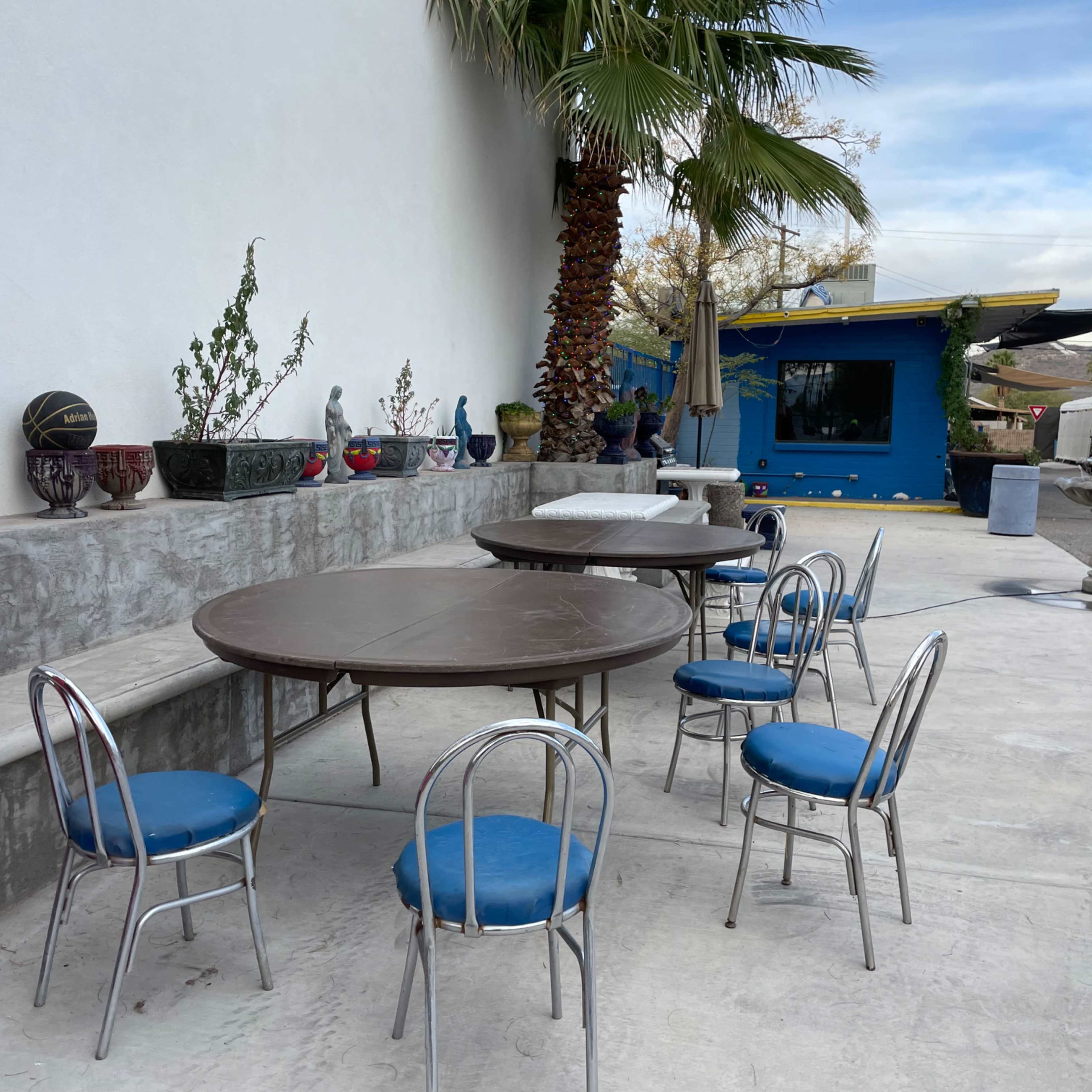 A seating area with two round tables and several blue chairs is set against a white wall, with a vending machine and decorative pots displayed nearby.