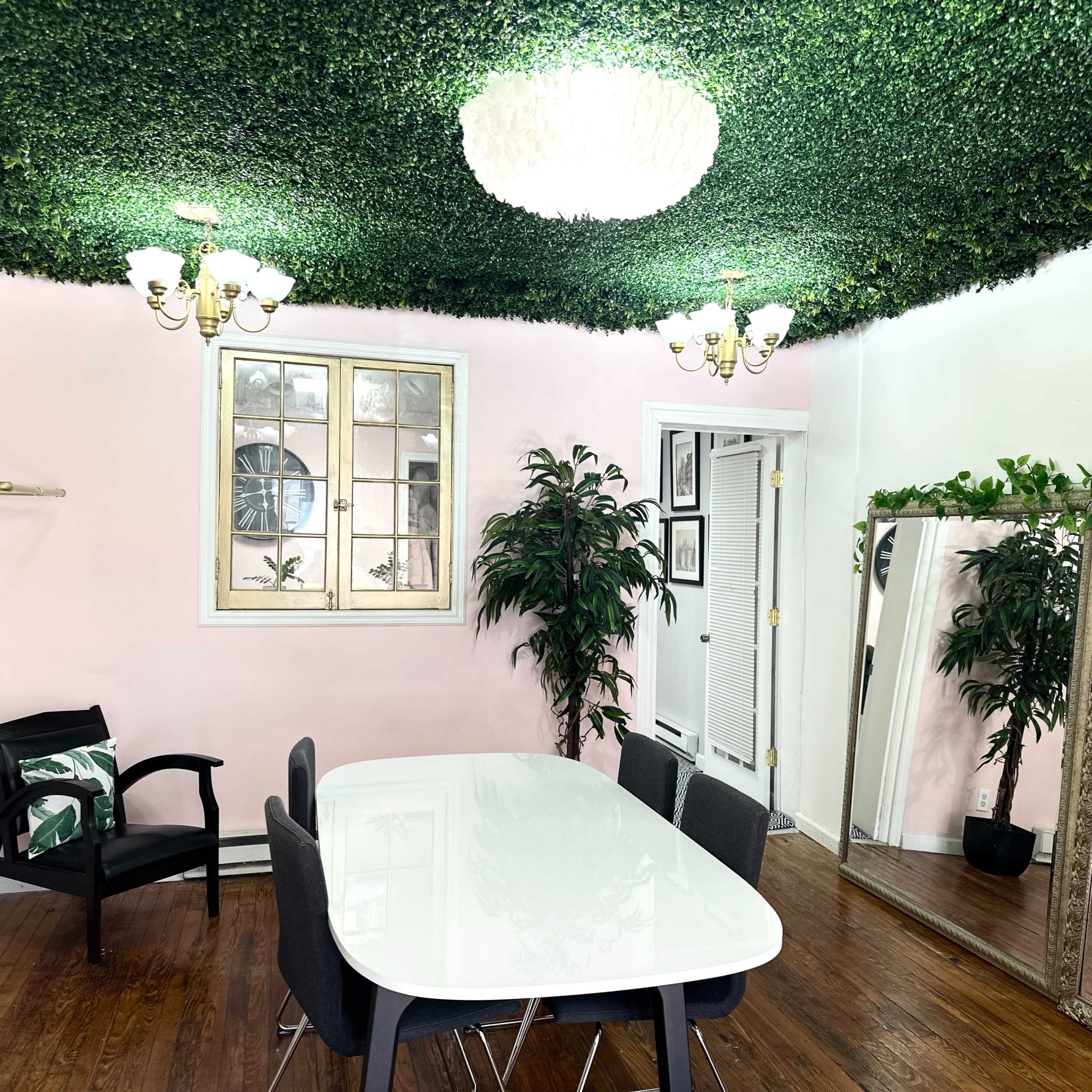 A brightly lit room featuring a white dining table surrounded by dark chairs, with a green, leafy ceiling, pink walls, and a large mirror.