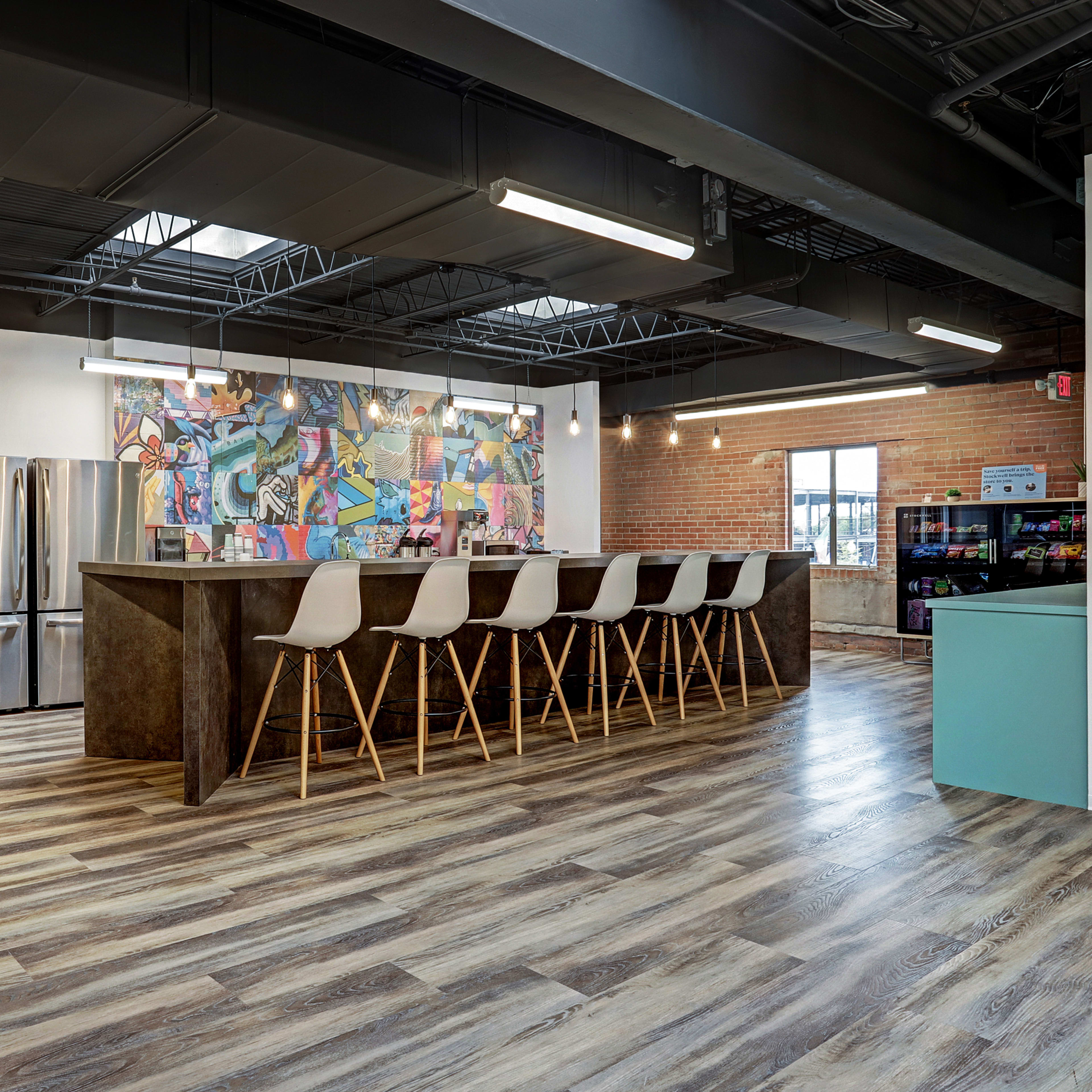 A modern office kitchen area with a bar counter and high stools, featuring a colorful mural on the wall and stainless steel appliances alongside a snack area.