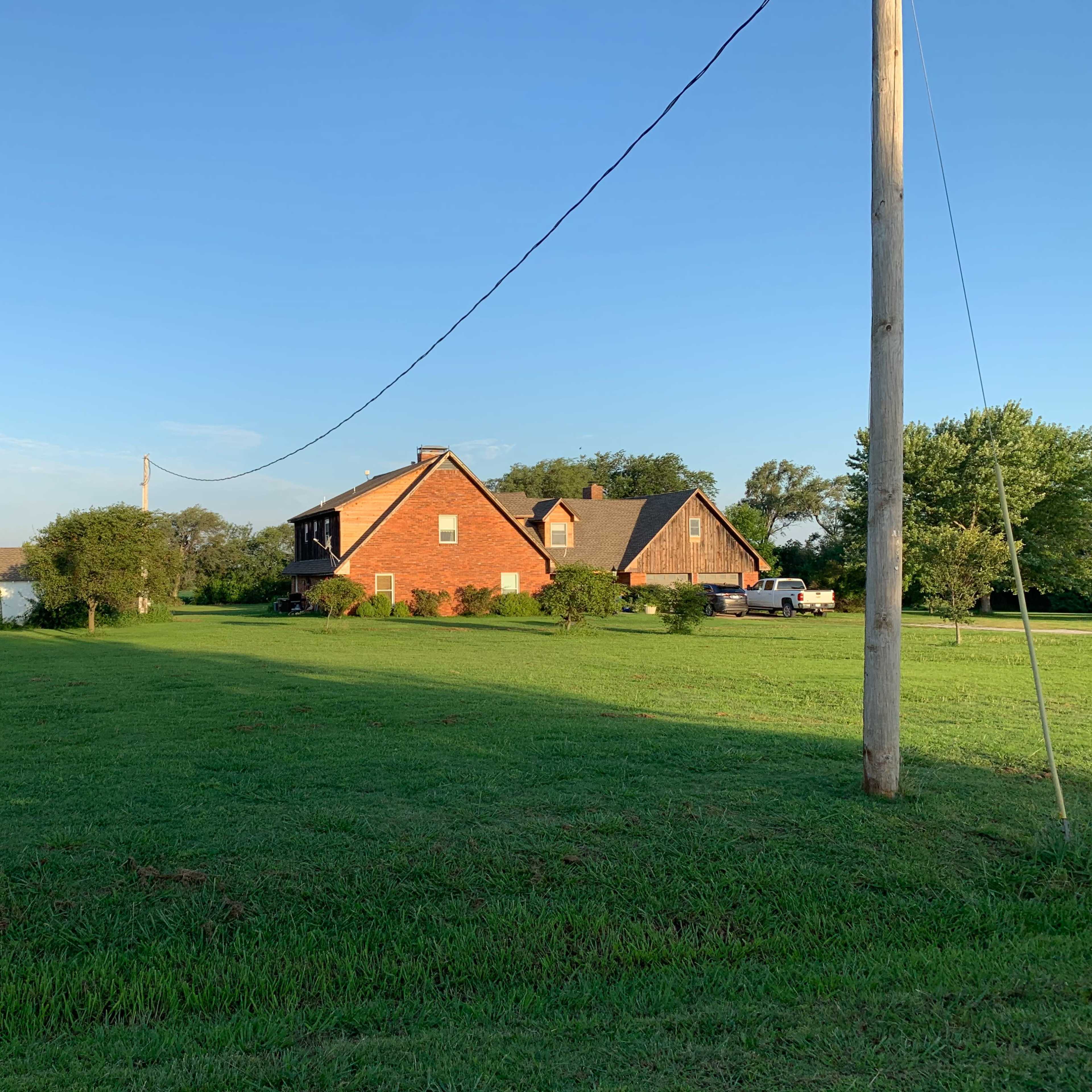 A red brick house with a sloped roof is set on a grassy lawn, surrounded by trees and utility poles under a clear blue sky.