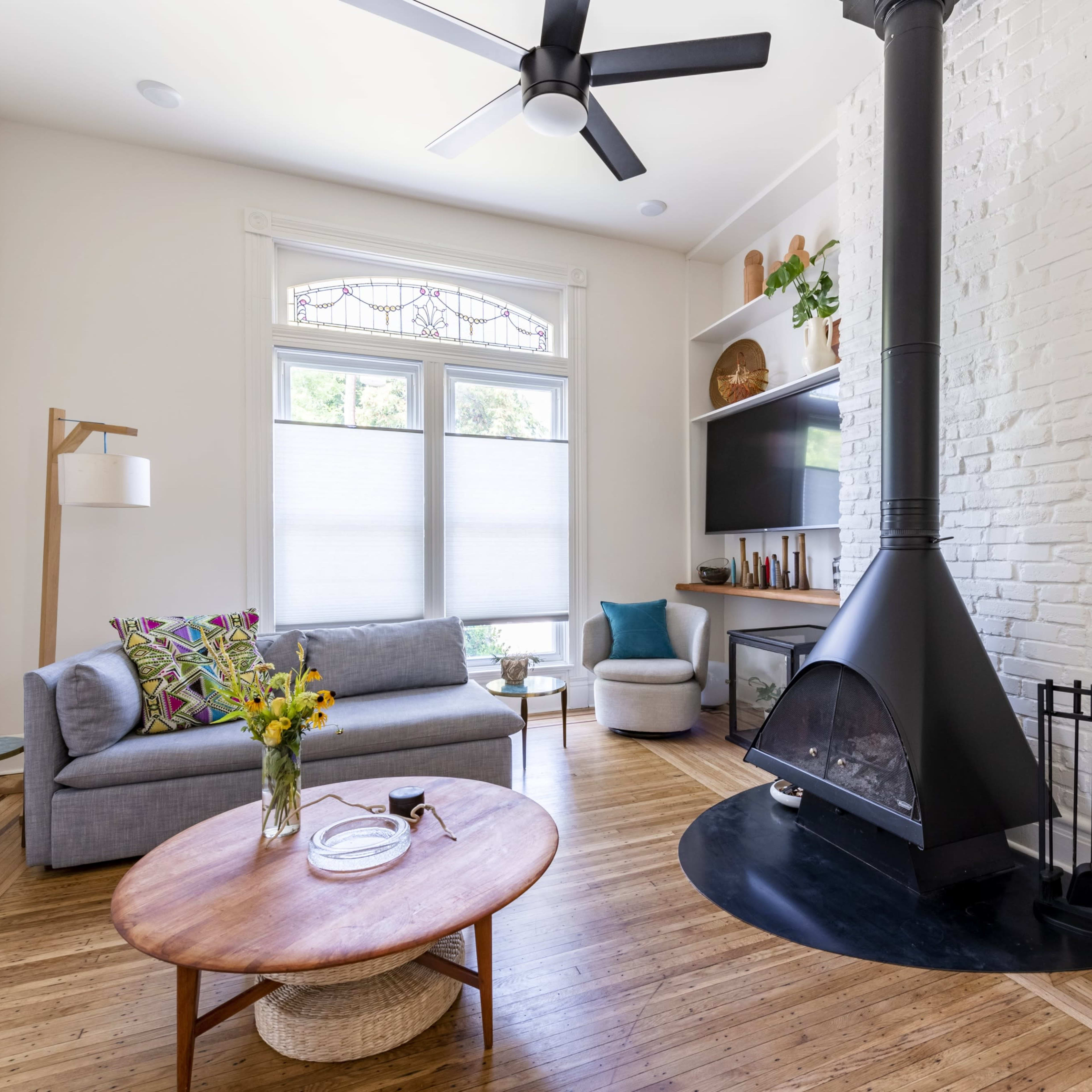 A modern living room featuring a gray sofa, a round wooden coffee table, and a black fireplace nestled against a white brick wall.