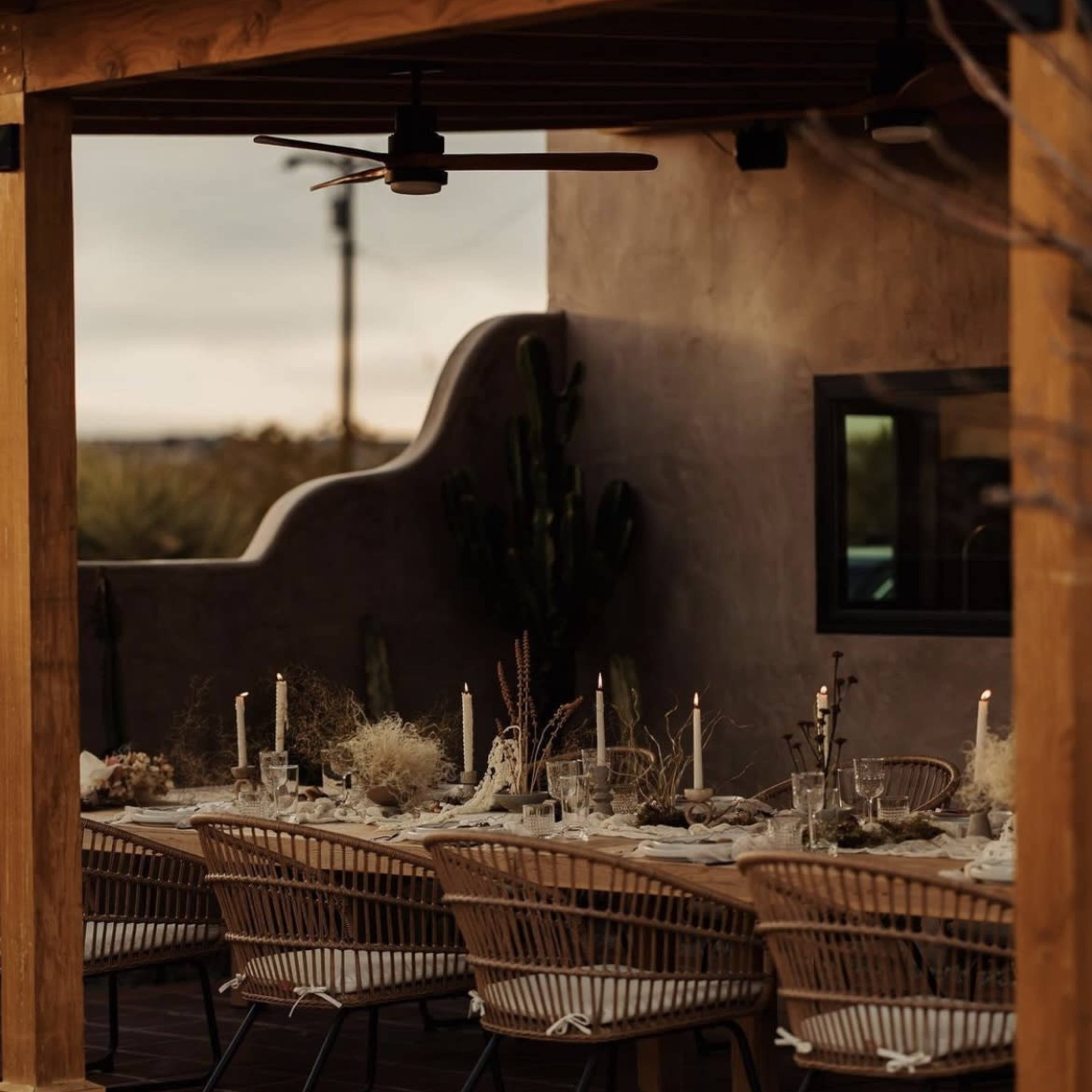 A long wooden dining table is set with candles and decorative elements under a covered patio surrounded by cacti.
