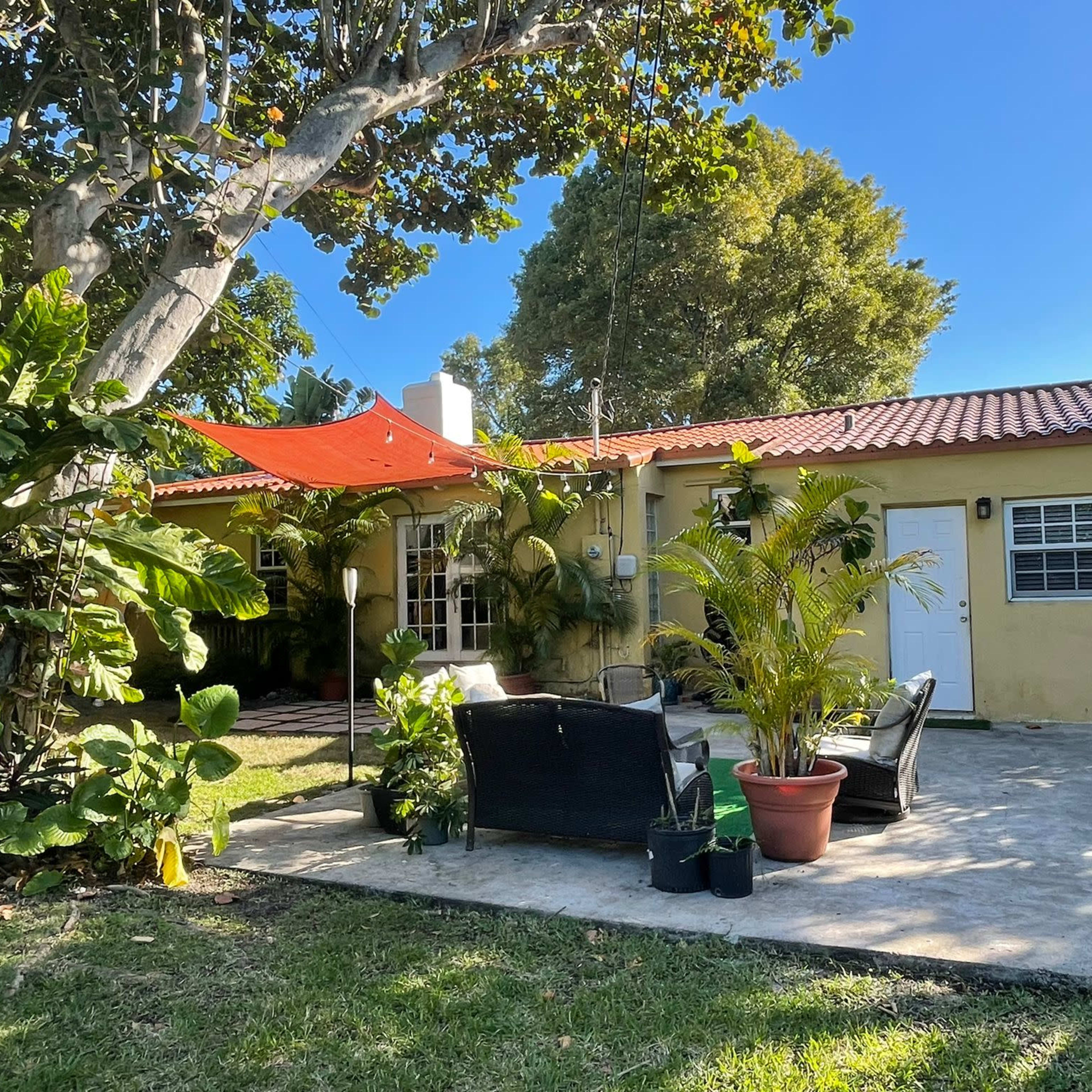 A small house with a red awning is surrounded by tropical plants and features a patio area with seating furniture.
