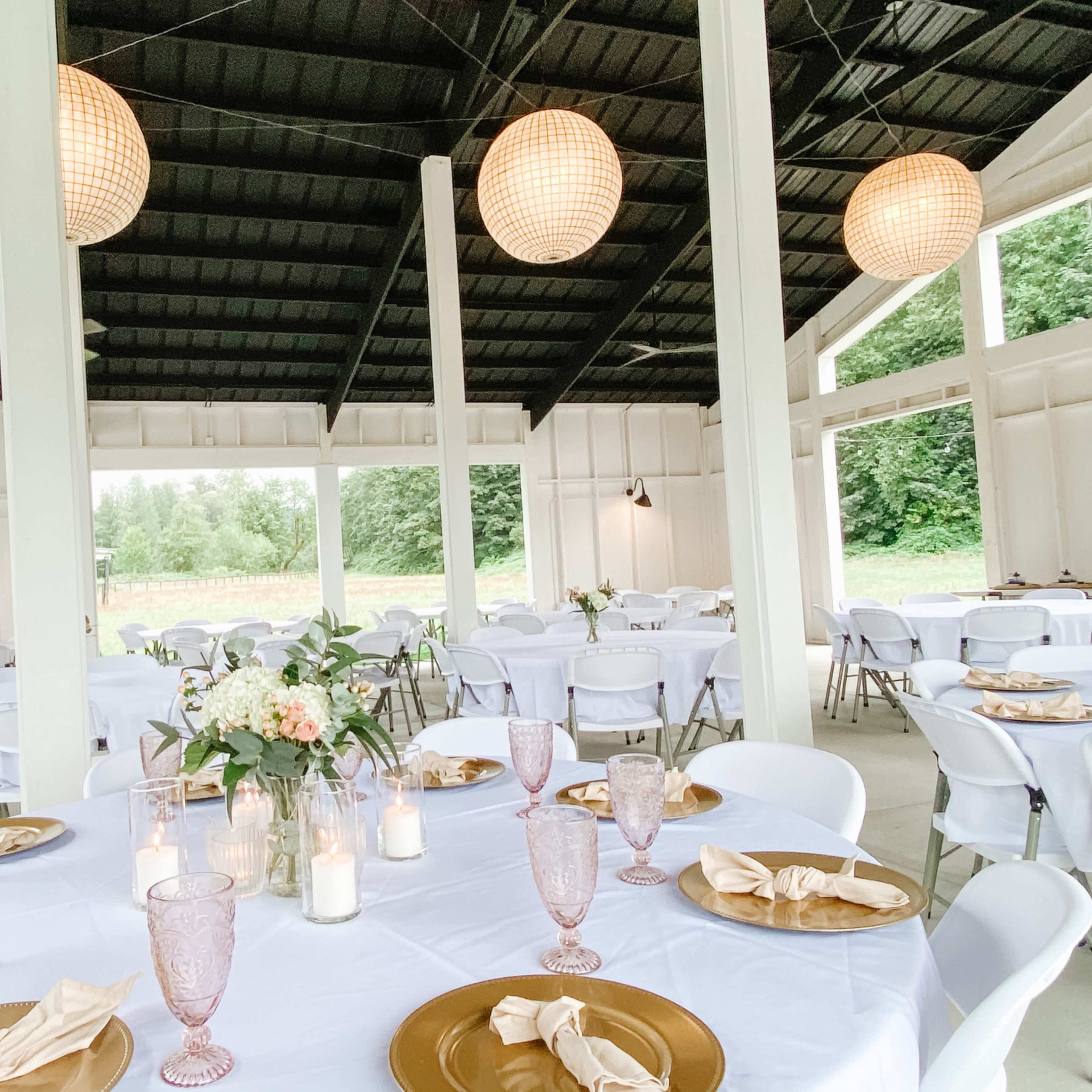 An indoor event space features white tables set with golden chargers, glassware, and floral centerpieces, illuminated by woven pendant lights.