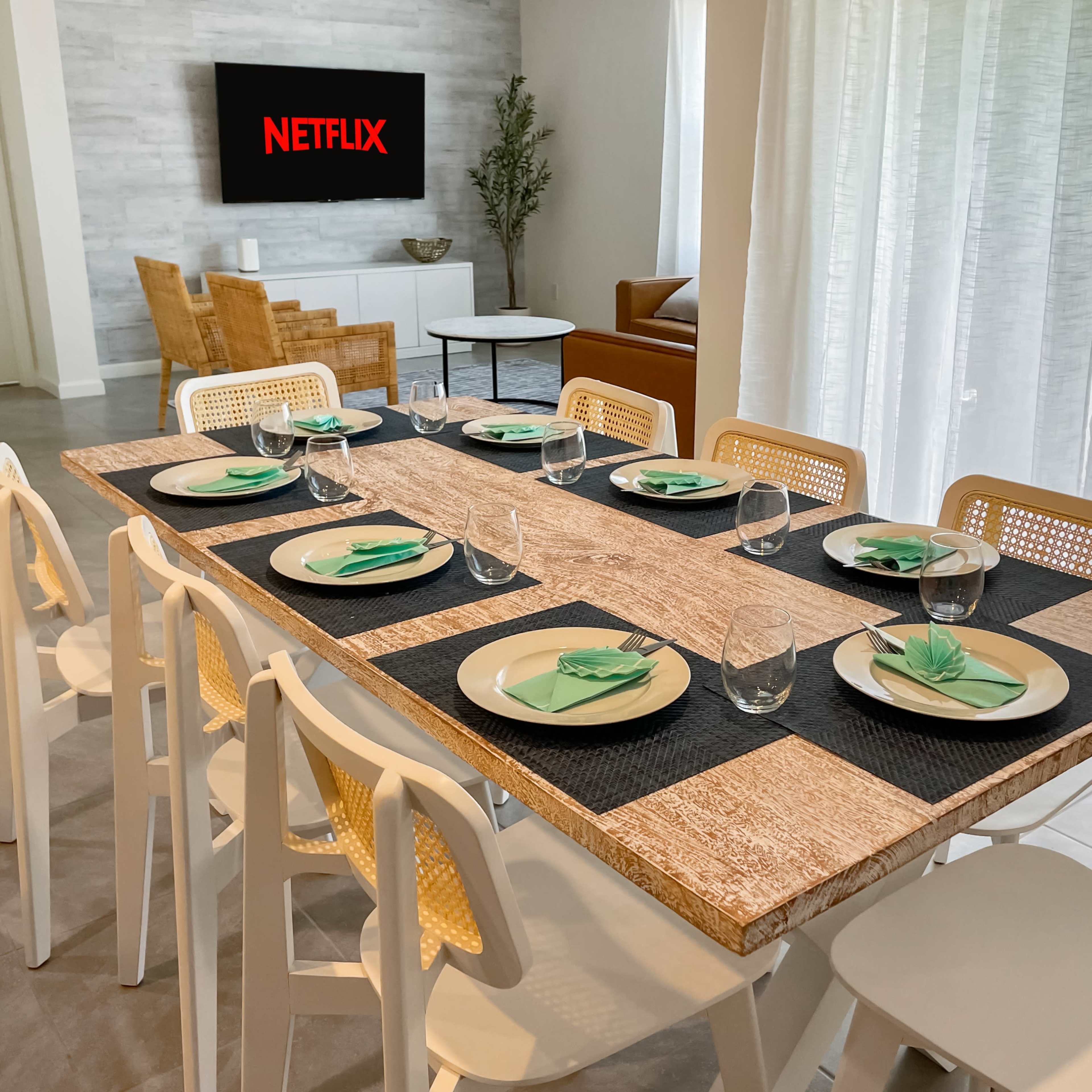A dining table is set for eight with plates and glasses, and a television displaying the Netflix logo is mounted on the wall in the background.