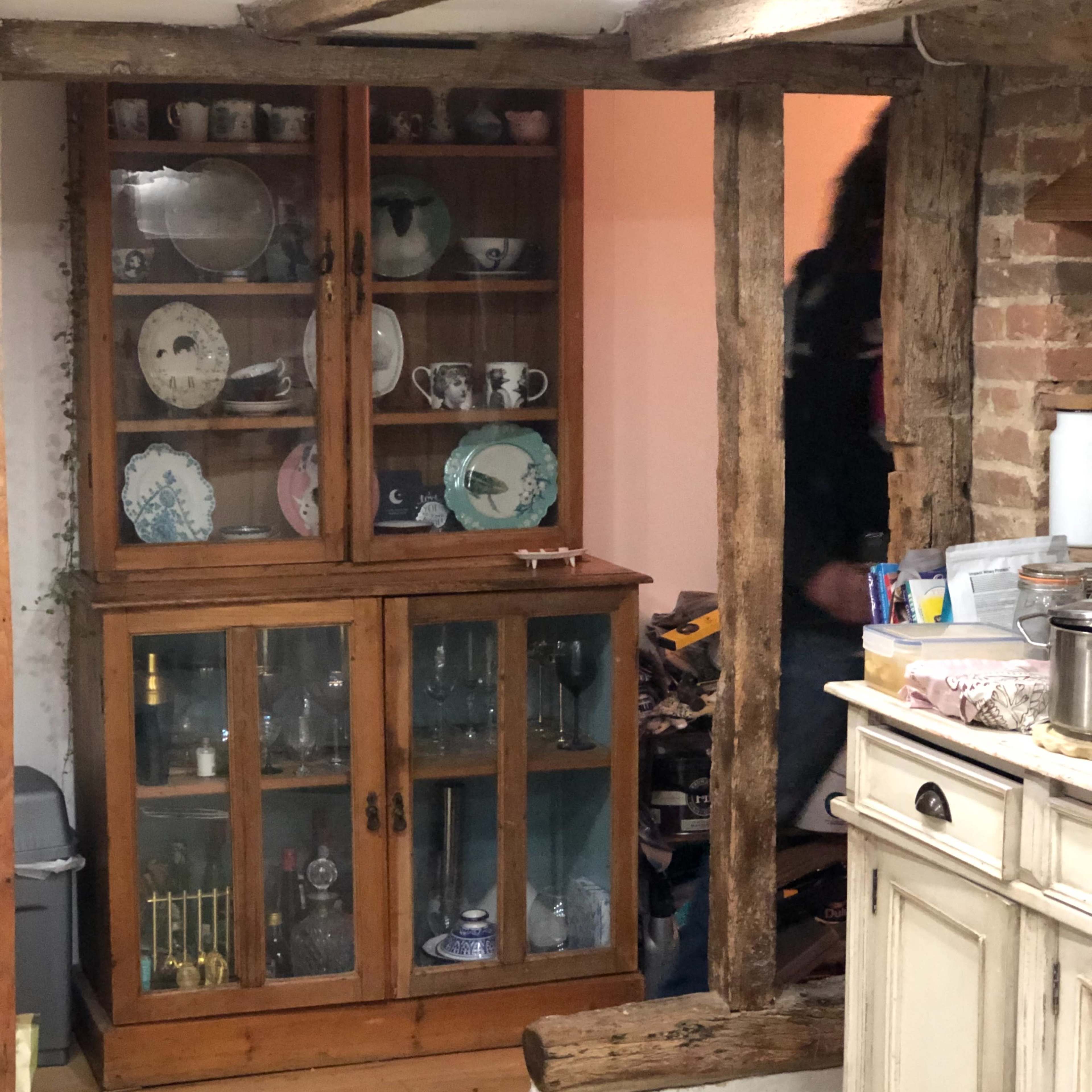 A kitchen area with a wooden display cabinet containing various plates and glassware, adjacent to exposed wooden beams and a partially visible person.