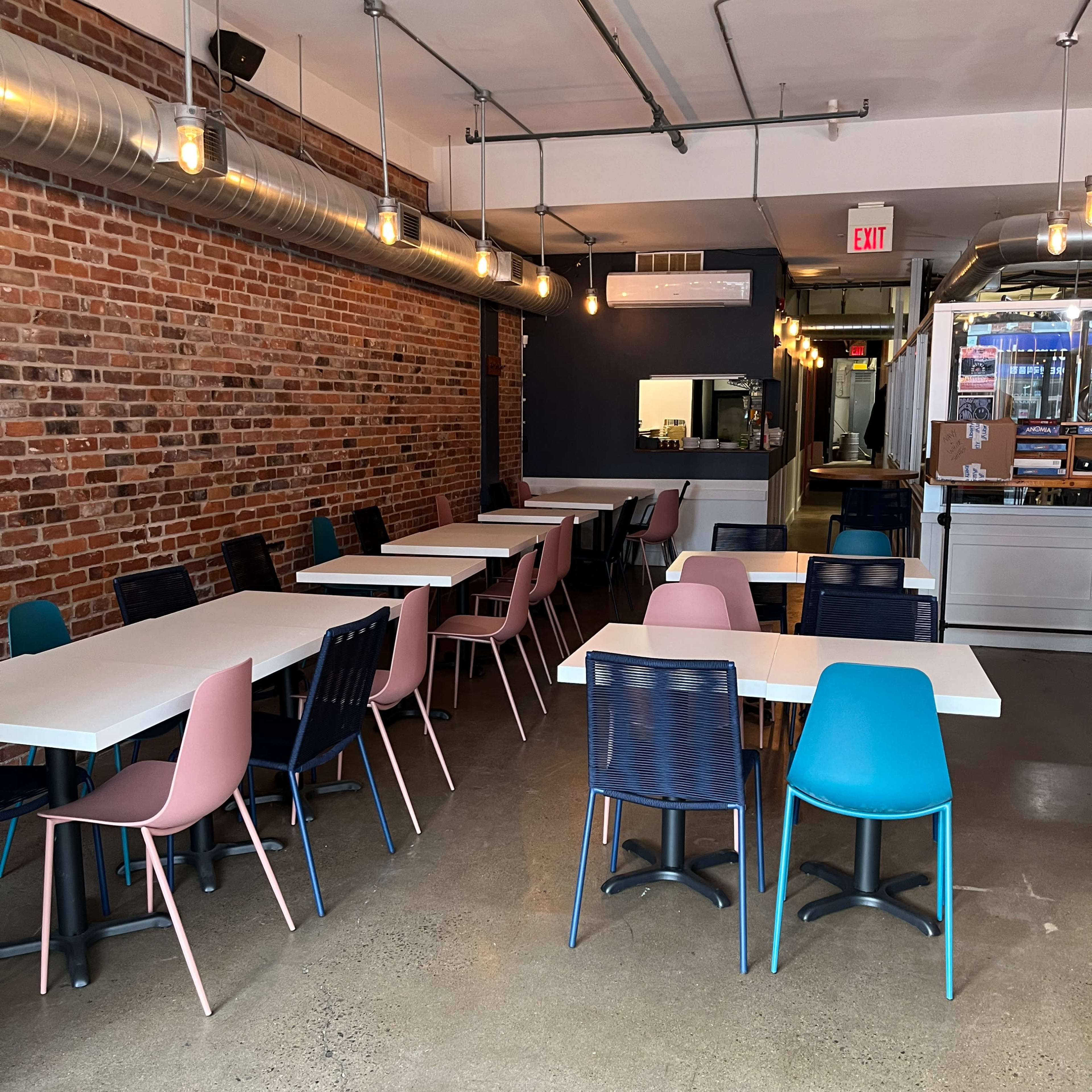 A spacious cafe with a mix of light pink, blue, and dark chairs arranged around white tables against a backdrop of exposed brick and industrial-style lighting.