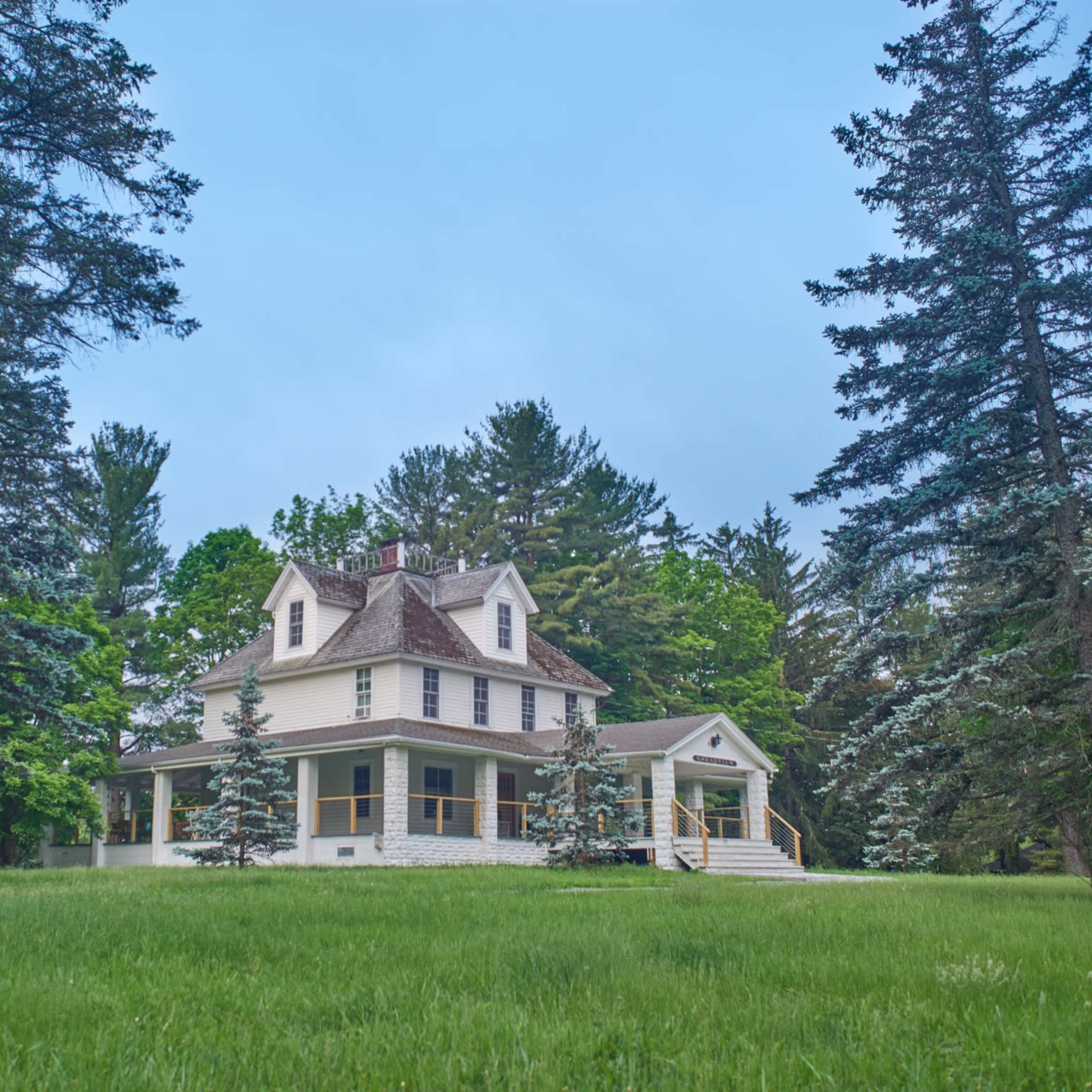 A large, two-story white house with a wraparound porch is surrounded by tall evergreen trees and well-maintained lawn.