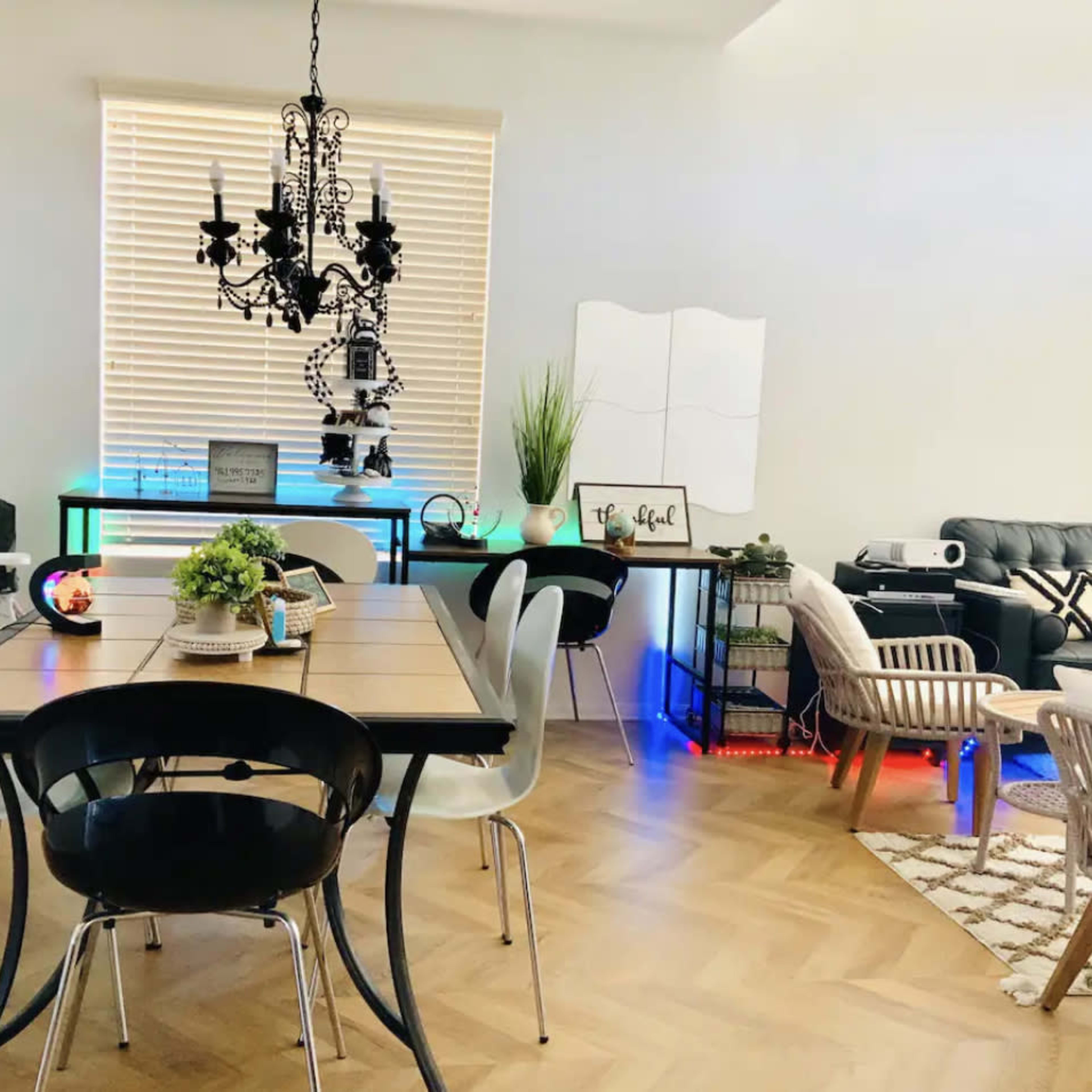 A dining area features a wooden table surrounded by black and white chairs, with a modern chandelier above and decorative plants on a side table.