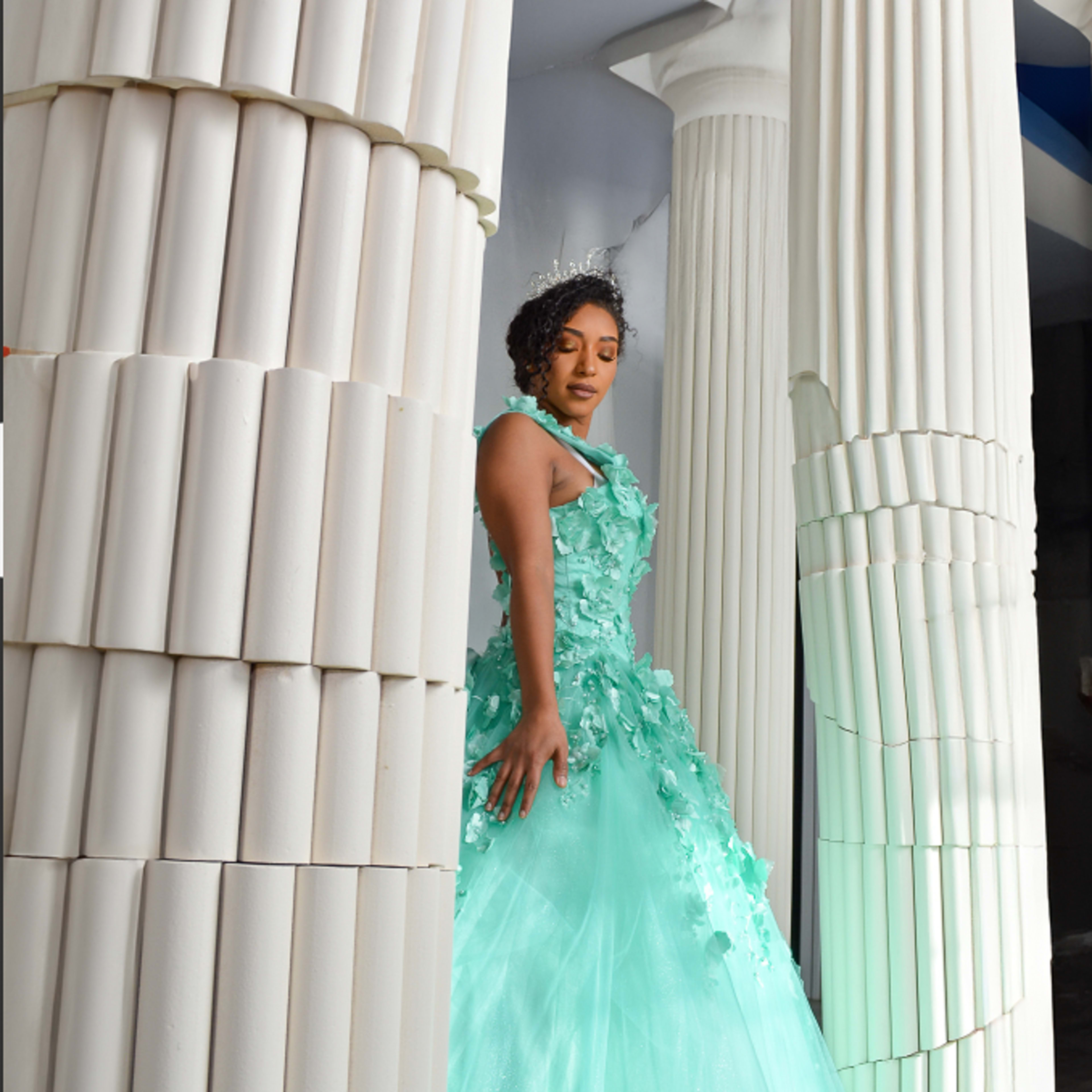 A woman in a mint green gown adorned with floral accents poses between two classical columns.