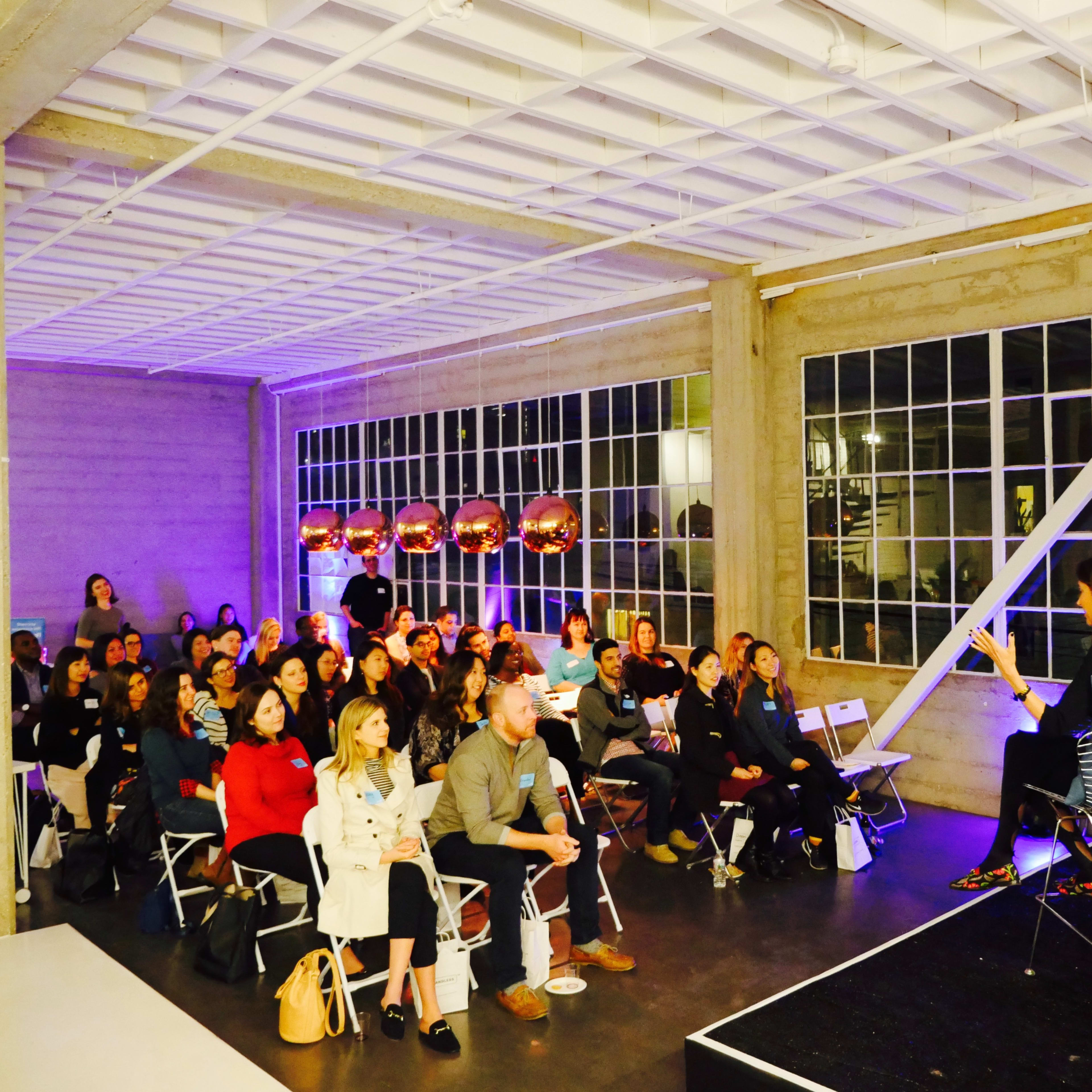 A large audience is seated in a spacious, well-lit venue, listening to speakers on a stage during an event.