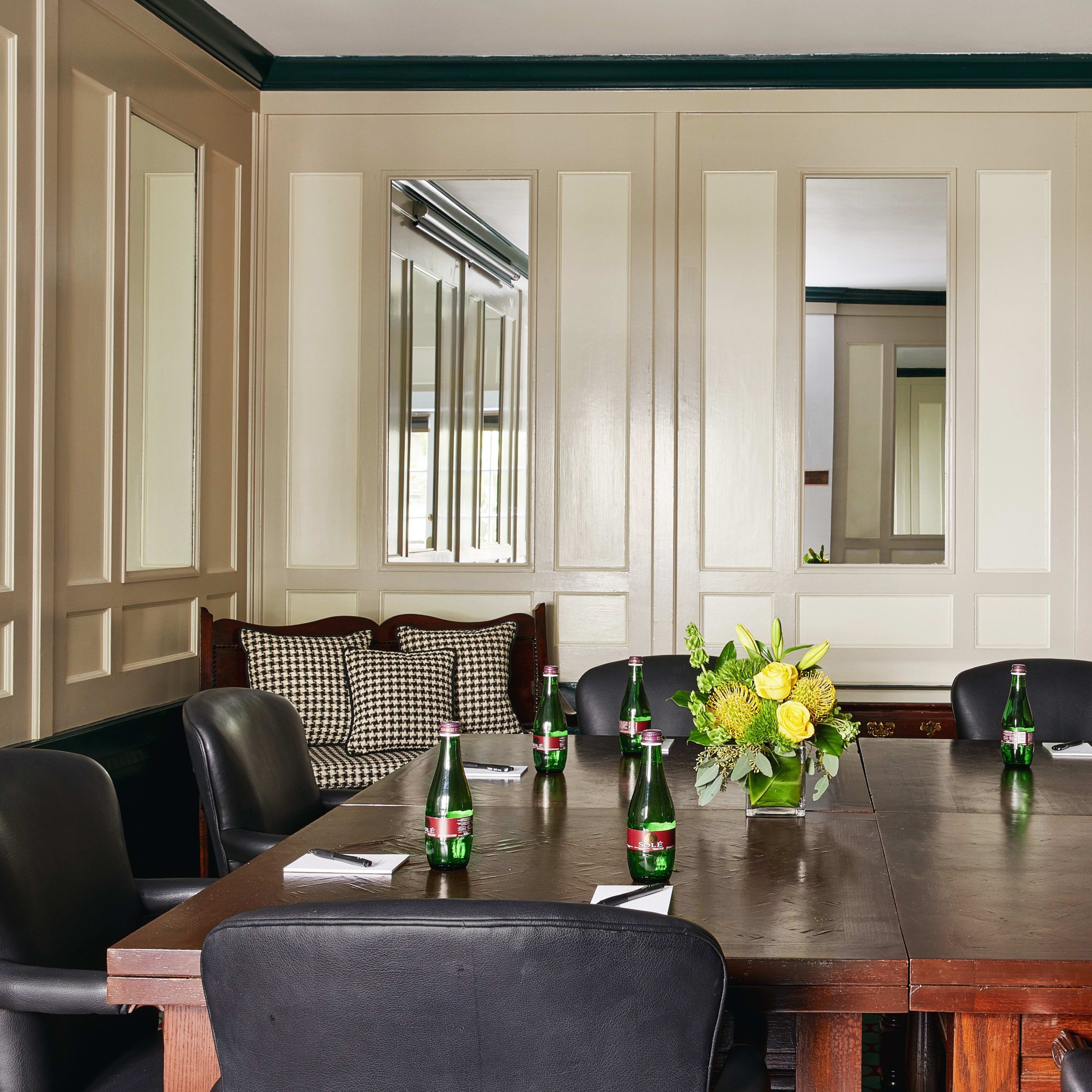 A conference room features a wooden table surrounded by black chairs, decorated with floral arrangements and bottles of sparkling water.