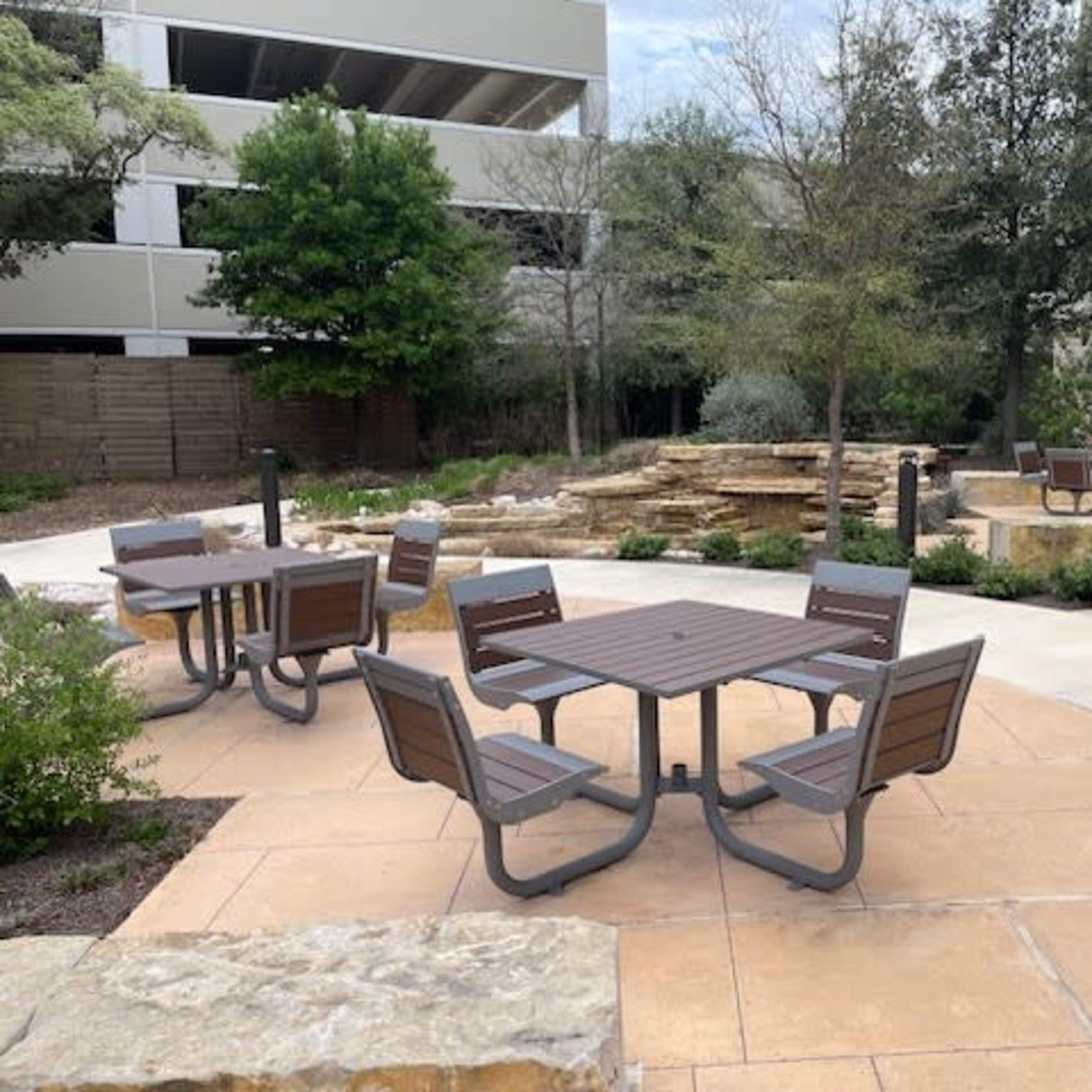 An outdoor seating area with several metal tables and chairs surrounded by landscaped greenery and a stone feature.