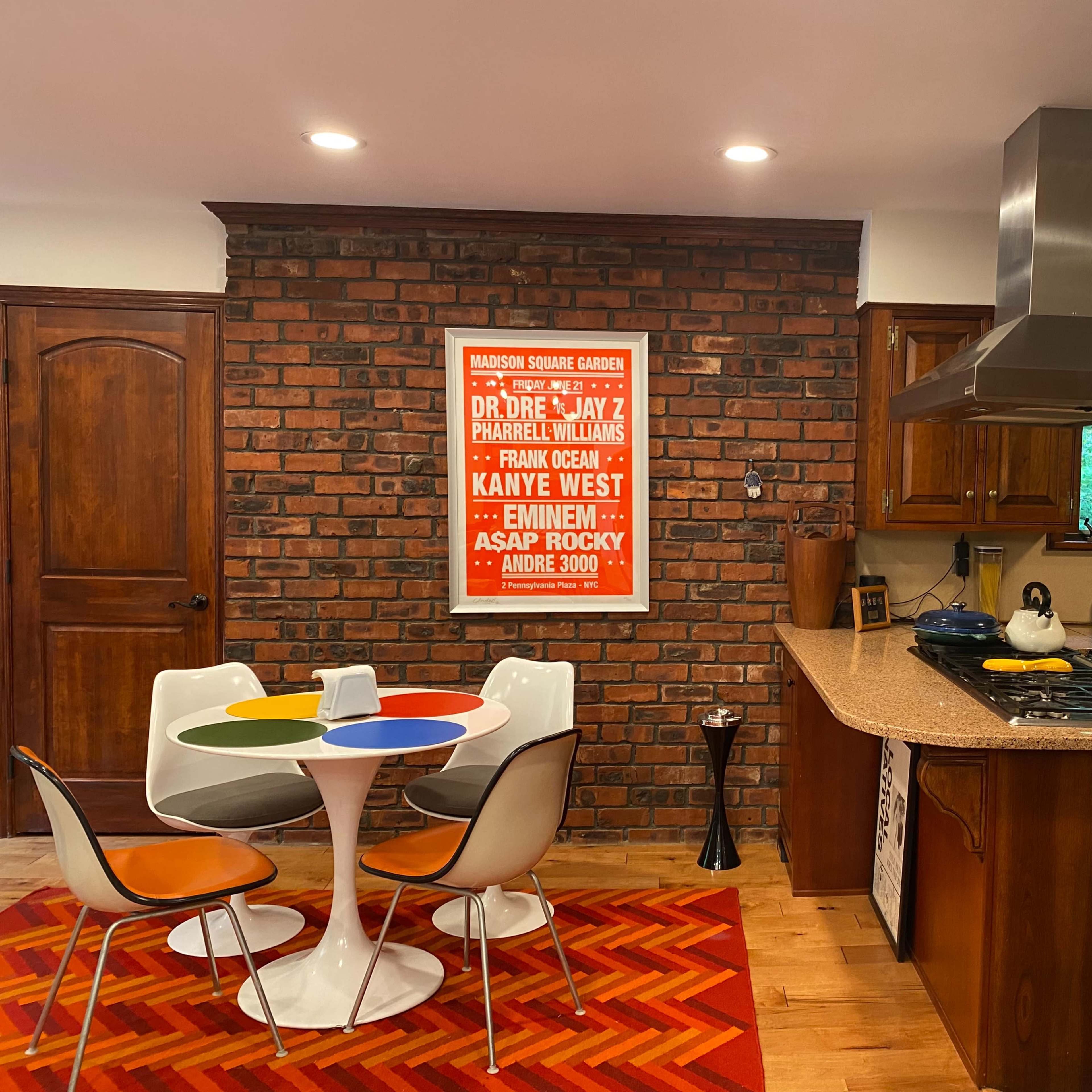 A modern kitchen features wooden cabinets, a granite countertop, and a round table with colorful chairs, highlighted by a large red poster on a brick wall.
