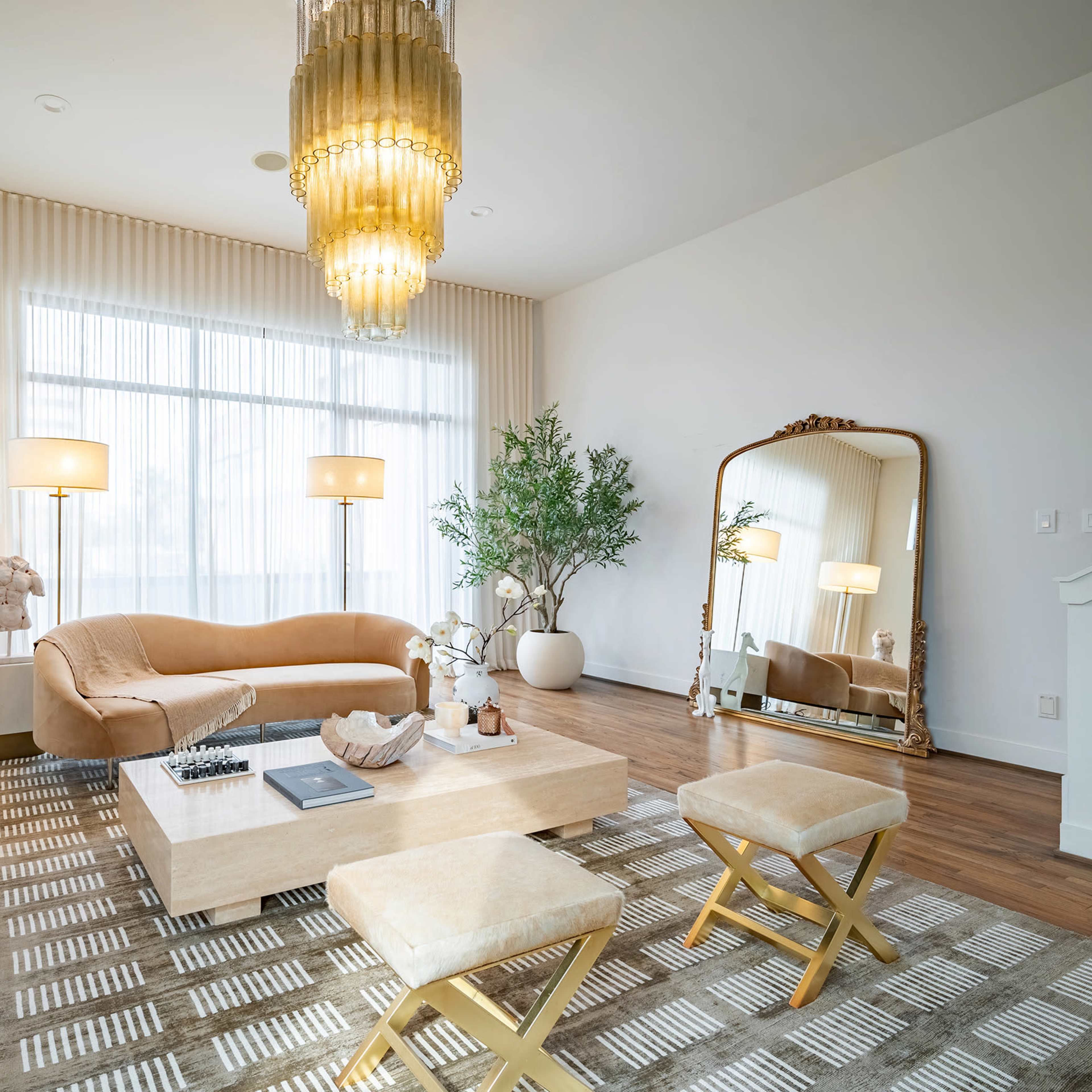 The living room features a light-colored sofa, a coffee table, decorative plants, and a large mirror under a chandelier, with tall windows letting in natural light.