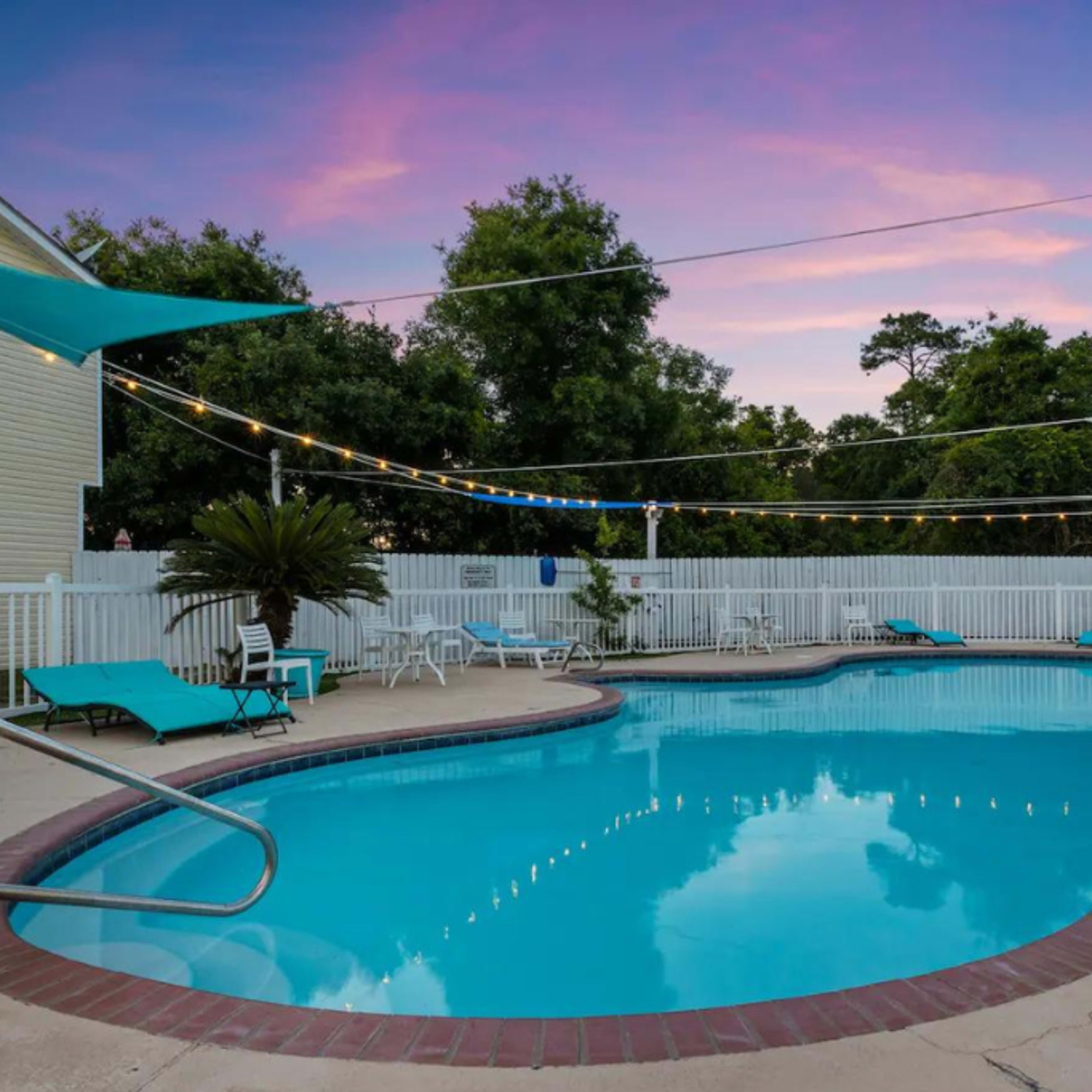 A swimming pool surrounded by lounge chairs, with illuminated string lights overhead and a wooden fence in the background.
